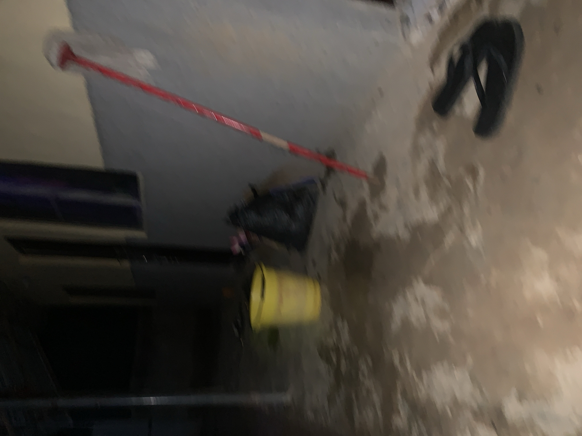 A blurred, tilted image depicts an interior scene. A red and white mop rests on a light-colored floor. Next to the mop is a black plastic bag, and a yellow bucket. To the right of the mop are black sandals. In the background on the left is a dark object. There are no people visible. The location is unknown, however the prompt suggests the location is Keffi, Nigeria. The image quality prevents a definitive assessment.