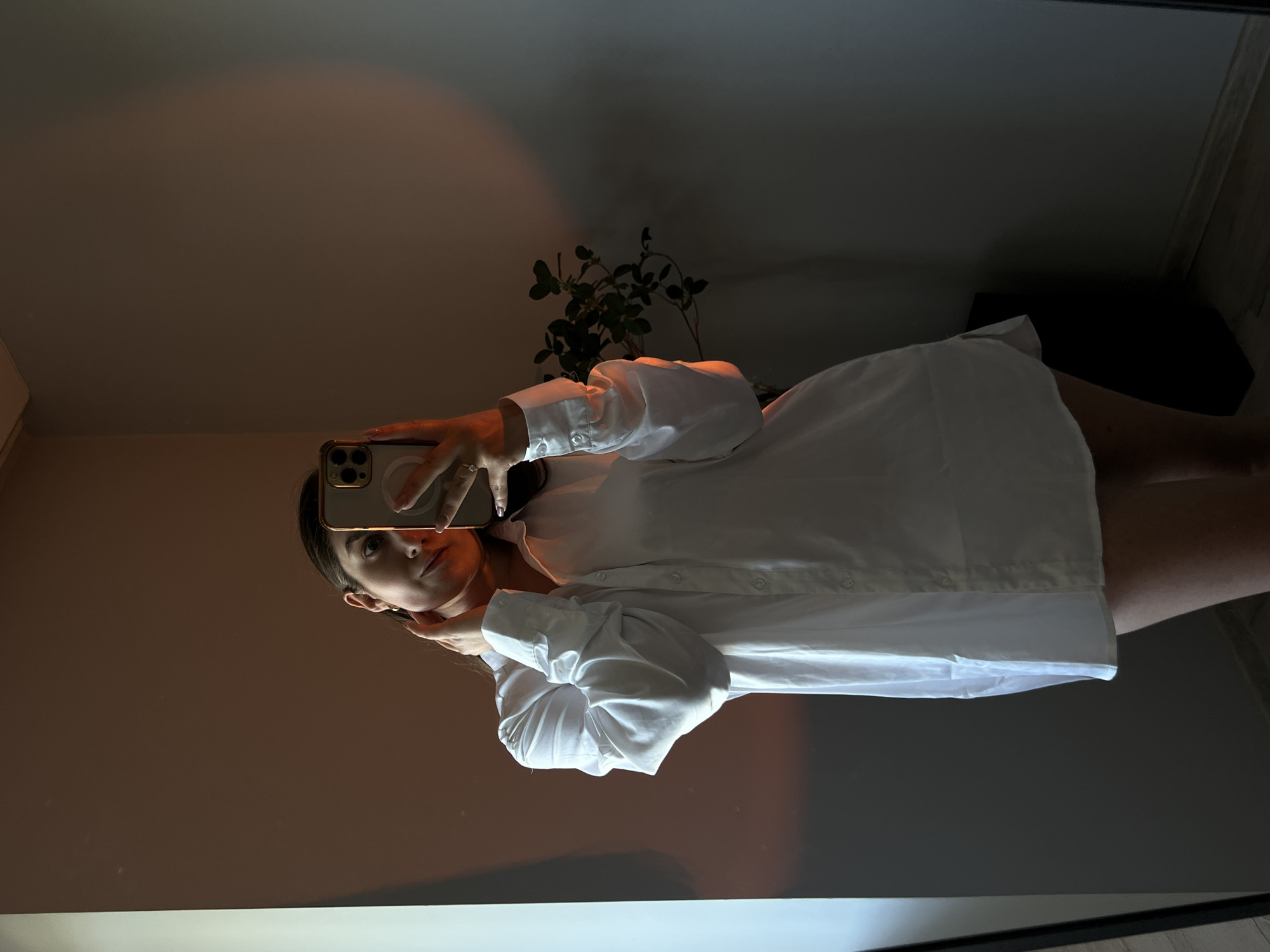 A woman in a white oversized shirt takes a selfie in a room with warm lighting. She is touching her neck with one hand, and holding her phone with the other. A plant is visible in the background. The setting appears to be a residential interior, possibly an apartment.