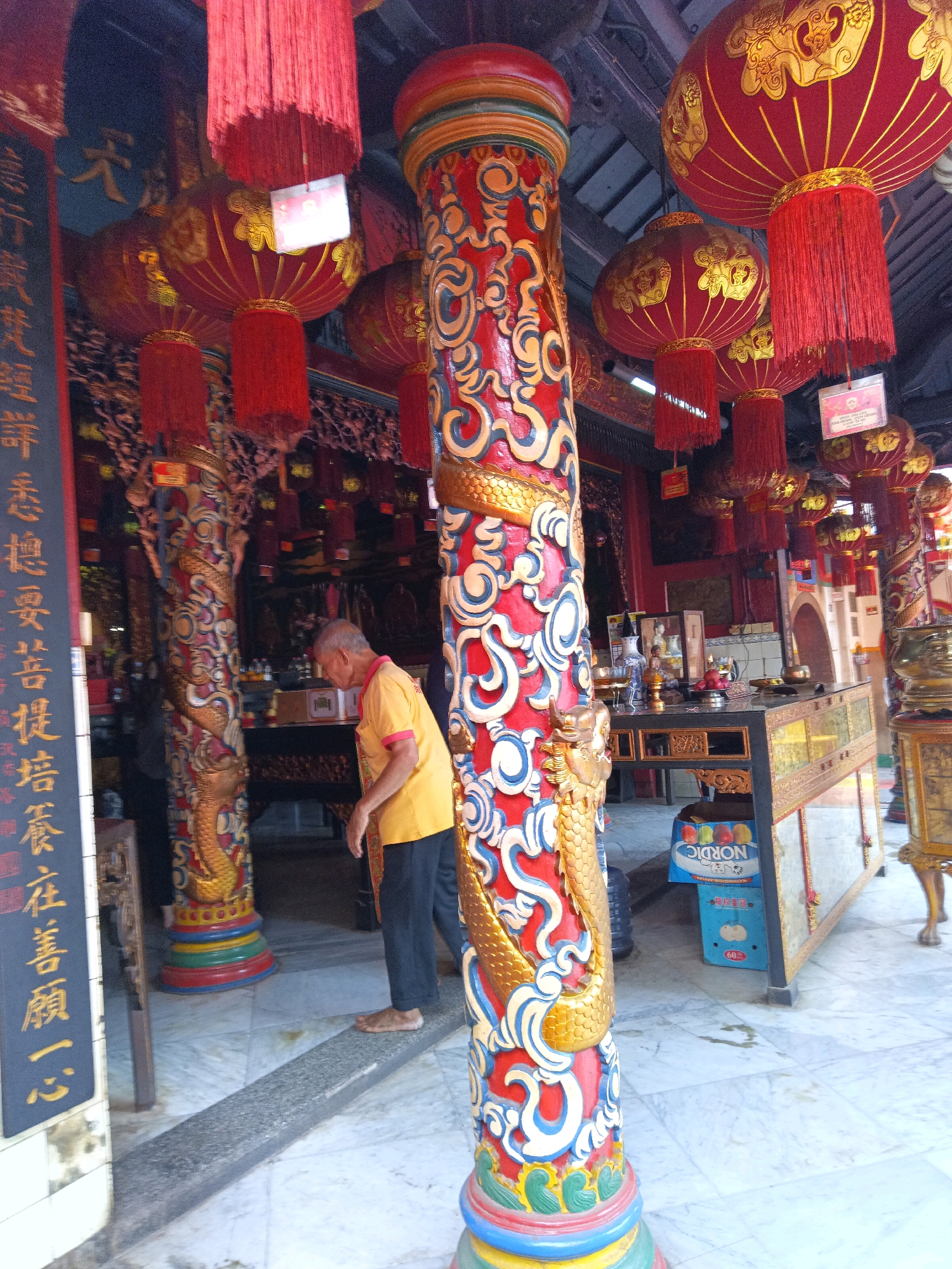 The image captures an indoor scene within a temple or religious establishment, likely in Tangerang, Indonesia, judging by the contextual information. The foreground is dominated by a vibrantly decorated pillar featuring a coiled golden dragon sculpture and intricate red, blue, and white scrollwork. Red lanterns with gold embellishments and fringe hang in the background, contributing to a festive and spiritual ambiance. An elderly man, wearing a yellow short-sleeved shirt and dark trousers, is partially visible in the middle ground, facing away from the camera and appearing to be engaged in an activity or contemplation. He is barefoot and stands on a raised stone platform. To the right of the man, a long, ornate table with glass panels and drawers is laden with various objects, including vases, figurines, and what appear to be offerings. A blue cardboard box labeled "NORDIC" is positioned on the floor beneath the table. The floor is tiled with a marble-like pattern. The overall scene is characterized by rich colors, detailed craftsmanship, and a serene atmosphere, indicative of a place of worship or cultural significance.
