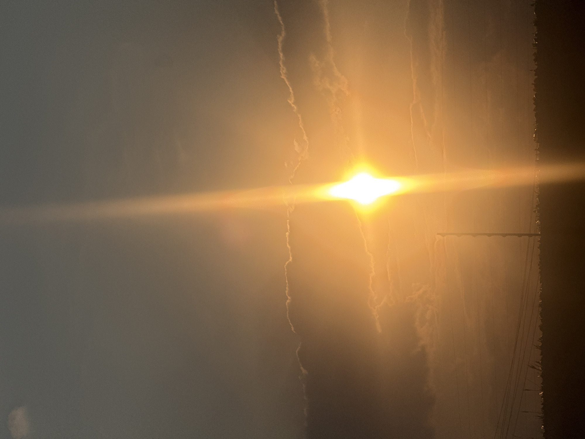 A sunlit sky is depicted in the image, with the sun positioned centrally and emitting a bright, horizontal glare. To the right of the sun, there are utility poles and lines. The atmosphere appears hazy or smoky. The location is Unknown, Unknown.