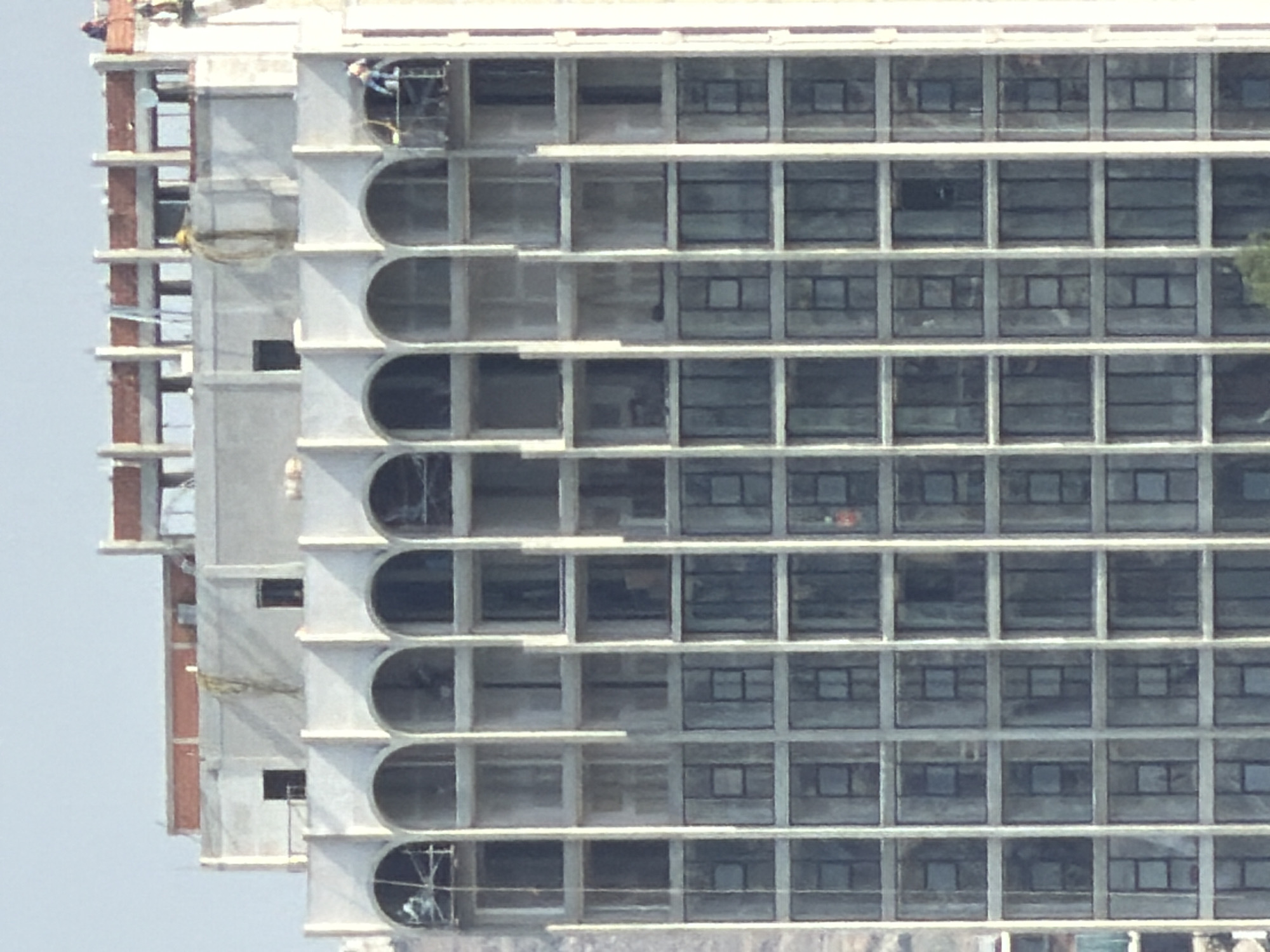 The image depicts the side of an unfinished high-rise building, likely under construction. The building features a series of rectangular window frames interspersed with sections of rounded arches. There are a few people visible, seemingly construction workers, located at the top and bottom of the exposed structure. The building has a partially constructed concrete and steel frame. The location of the image is Santa Clara Ocoyucan, Mexico.