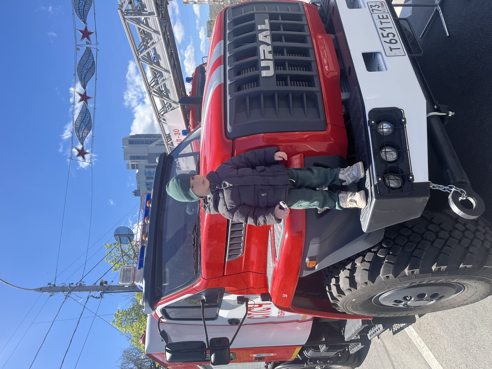 A young boy stands on the front of a red and white Ural fire truck. He is wearing a dark jacket, green pants, a green hat, and light-colored shoes. The fire truck's license plate reads "T651TE73RUS". The "URAL" logo is prominently displayed on the front of the truck. The background shows a blue sky with clouds and a building in the distance. A decorative light fixture with stars is visible in the upper left corner. This photo was likely taken in the city of Ulyanovsk, Russia.