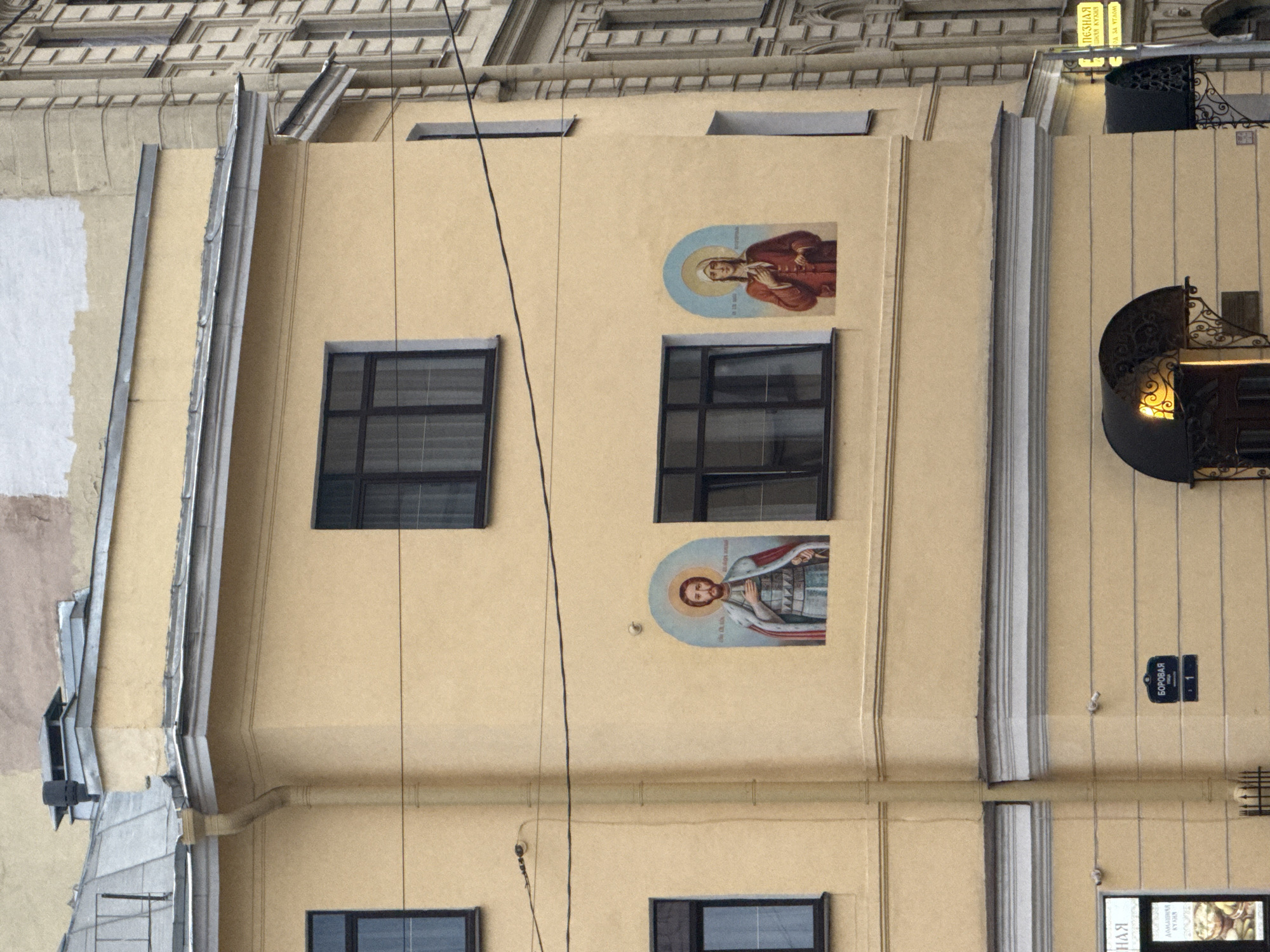 Here's a description of the image:
The image shows the facade of a building in Saint Petersburg, Russia. The building is light yellow in color and has two rows of windows with dark frames. On the building's facade, between the windows, are two paintings of religious figures depicted in iconographic style. At the top right of the building is a sign with Cyrillic text and an elaborate wrought-iron balcony. Lower on the right side of the building, a street sign labeled "БОРОВАЯ" (Borovaya) with the number "1" is visible. Below the sign is the corner of a building with a sign written in Russian. In the top left of the image are parts of the upper levels of a neighboring building. A black wire runs vertically down the building's facade.