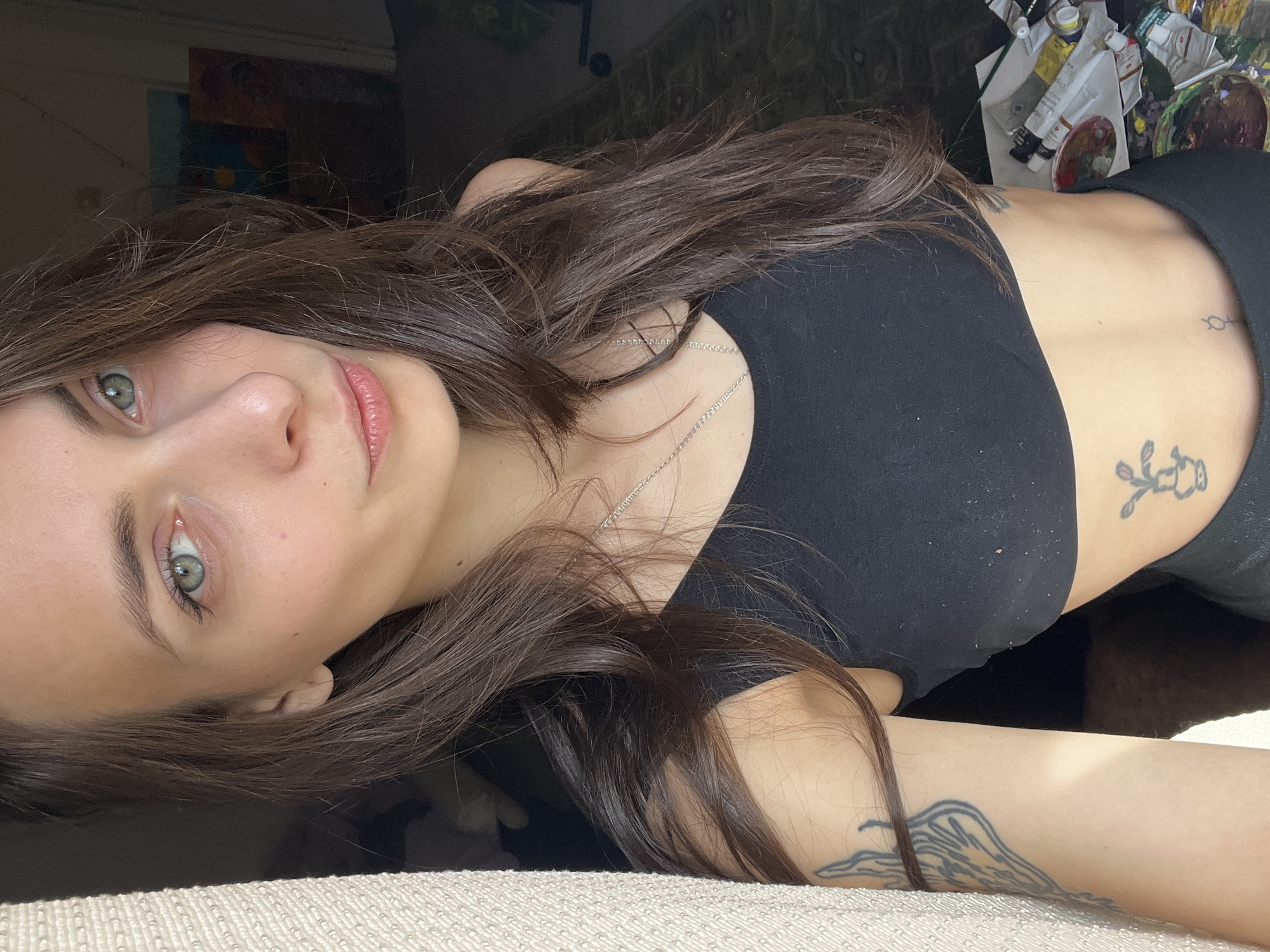 A young woman lies on a light-colored textured surface. She is wearing a black crop top and dark pants. She has long, dark brown hair and light green eyes. A silver necklace is visible on her neck. Tattoos are seen on her stomach and arm. In the background, there is a cluttered area with art supplies and a window. The image does not contain sufficient information to identify the location as Krasnoyarsk, Russia.