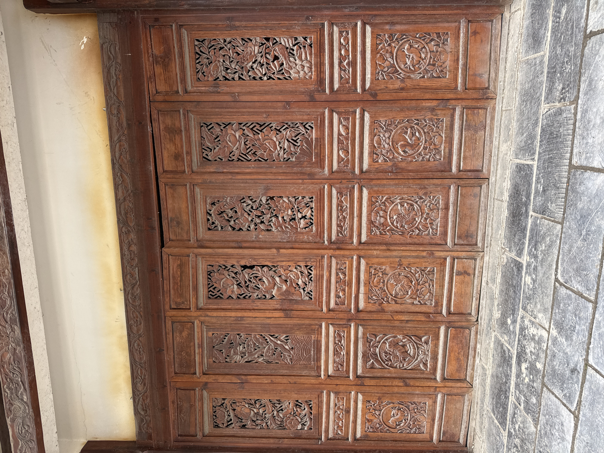 The image displays a detailed view of an ornate, dark brown wooden facade, likely a building entrance, located in Wulingyuan, China. The facade comprises six vertical panels, each segmenting into multiple carved sections.
The upper portion of each panel features a recessed rectangular area with a lattice-like background behind elaborate, deep relief wood carvings. These carvings depict complex arrangements of birds, including what appear to be peacocks or phoenixes, amidst dense floral and foliage motifs, specifically identifiable as peonies in some instances. Below these, smaller horizontal panels are carved with continuous scrollwork or floral patterns. The lower part of each main panel showcases a prominent circular carving, each containing specific avian figures, such as swans or ducks, set against a floral backdrop. The lowest section of each panel consists of solid, uncarved rectangular wooden elements extending to the ground.
Above the wooden panels, a robust horizontal wooden lintel, also intricately carved with scrollwork, spans the facade's width. Above this lintel, a cream-colored wall section is visible, presenting minor surface cracks and distinct yellowish-brown discoloration or staining along its bottom edge, suggesting environmental exposure or moisture. A larger, darker horizontal wooden beam with additional carvings runs along the very top of the visible frame. The ground directly in front of the facade is paved with large, dark grey, rectangular stone slabs, exhibiting variations in texture and surface wear. No individuals or discernible activities are present in the scene, which captures a static architectural feature.