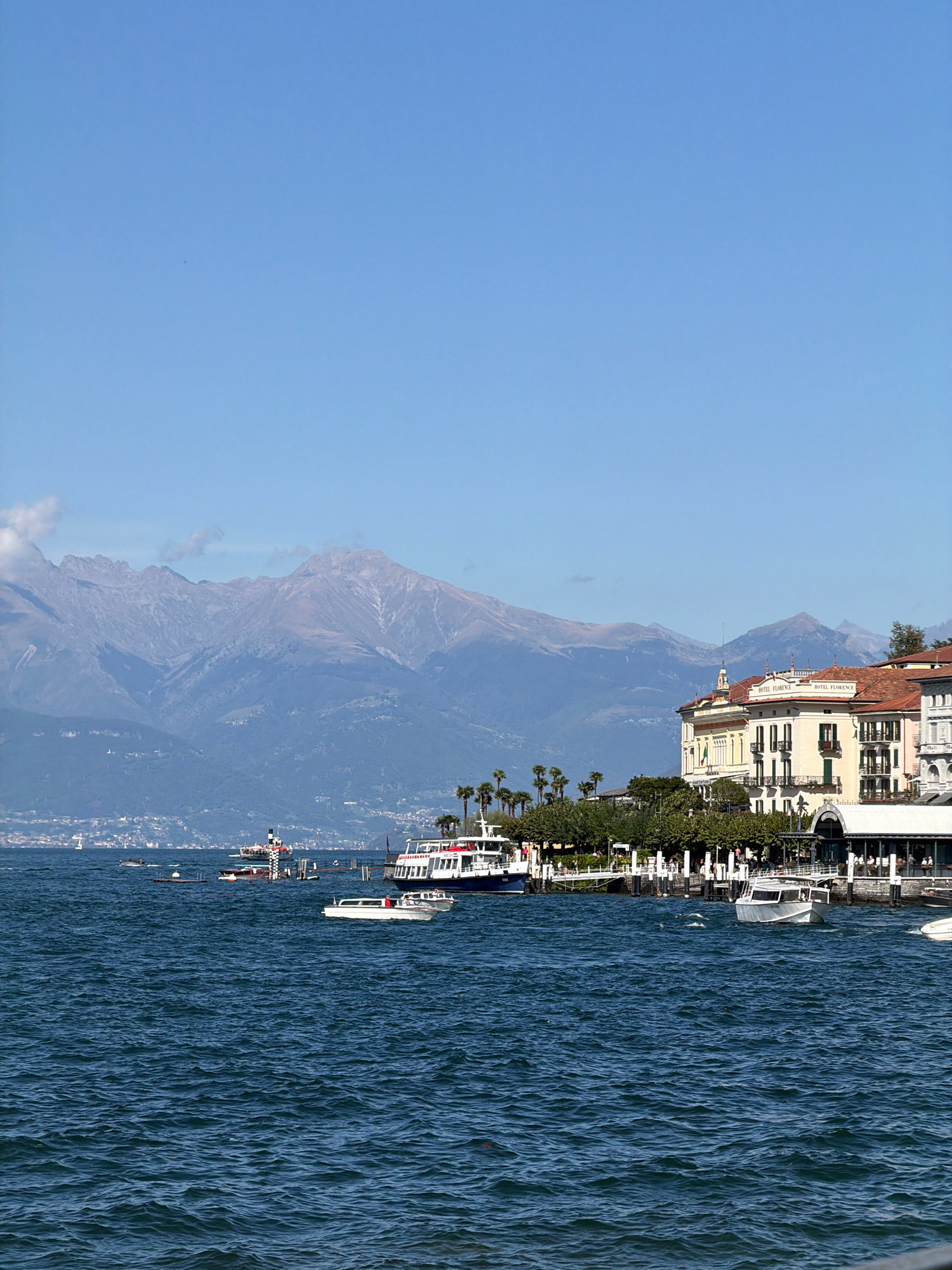 The image captures a scenic view of a lake with a backdrop of mountains. In the foreground, the deep blue water of the lake is agitated by ripples, indicating a breezy day. Several boats are visible on the water, including a white motorboat actively moving, and larger vessels docked along the shore. The middle ground features a waterfront promenade lined with trees and buildings. Prominent among these is a multi-story building with a red tiled roof, bearing the inscription "HOTEL FLORENCE." The building has several balconies with green shutters and a large arched entrance on the ground floor. Behind the waterfront structures, a range of imposing mountains forms the background, their peaks somewhat softened by atmospheric haze and possibly snow. The sky above is a clear, pale blue. The scene appears to be a busy tourist area, likely a lakeside resort town. While the prompt mentions "San Giovanni, Italy," this particular image does not contain any specific visual cues or landmarks that definitively place it in San Giovanni. The architecture and landscape are more broadly suggestive of a popular Italian lake district location.
