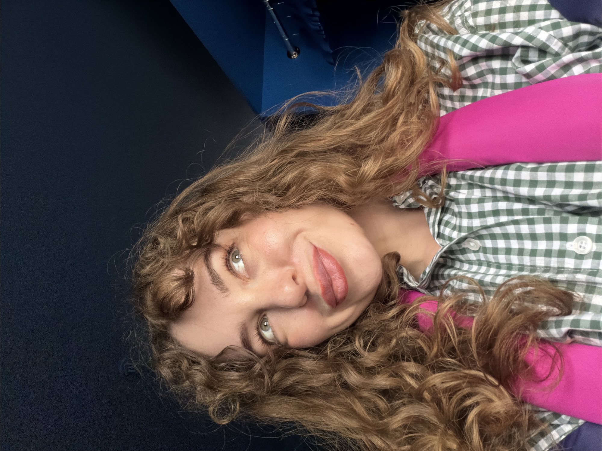 A woman with curly blonde hair and green eyes is lying down. She is wearing a green and white checkered shirt over a bright pink top. The background is a dark blue color. The photo appears to be taken indoors, possibly in a room with a dark blue wall. The location in the city of Moscow, Russia, cannot be determined from the image.