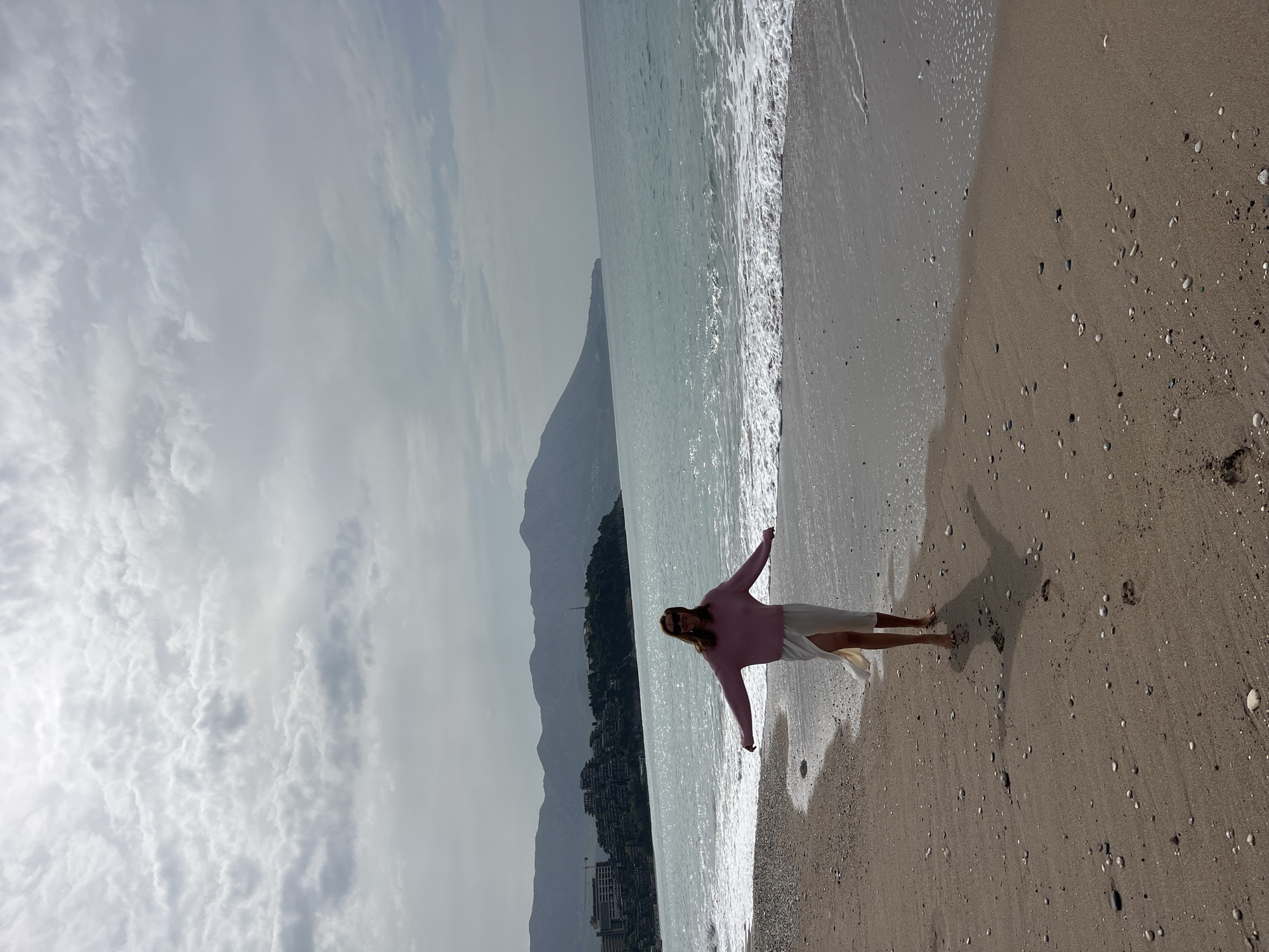 A woman stands on a beach with her arms outstretched. She wears a pink sweater and a white skirt. Behind her is the Adriatic Sea. In the background is a mountain with a city built at its base. The city is Budva, Montenegro. The sky is overcast. The beach is sandy with scattered pebbles.
