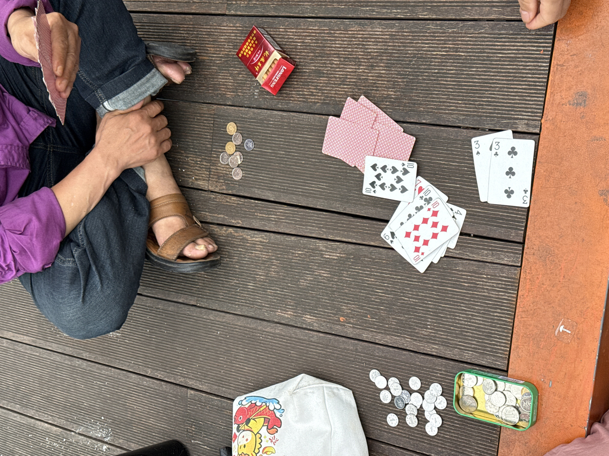 Observed in Nan'an District, China: An elevated perspective captures a group engaged in a card game on a wooden surface, presumably an outdoor terrace or deck. One individual is visible, seated, holding cards in hand, wearing jeans and a purple shirt. Coins are scattered nearby along with a white bag, a red matchbox, a green coin tin, and multiple playing cards scattered across the surface. The wood appears to be a part of a deck.