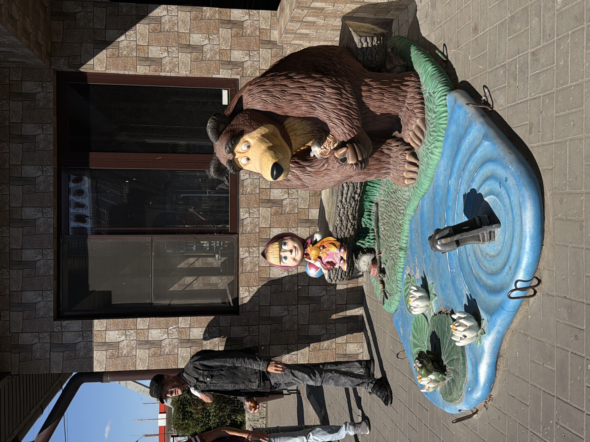 The image depicts an outdoor scene featuring several decorative figures and two individuals. The foreground is dominated by a blue, water-themed decorative base with several white water lilies and a green frog sculpture. To the right of this base is a large, brown, cartoonish bear sculpture, seemingly holding a small fish. Behind the bear, and to the left of it, is a smaller sculpture of a girl with blonde hair and a pink dress, holding a blue ball and a yellow toy horse. The sculptures are placed on a green, grass-like base. In the background, a man wearing a dark denim jacket, dark pants, and a cap stands facing to the left, holding a small, round, orange object in his right hand. Another person, wearing light blue jeans and gray shoes, is partially visible to the far left of the frame. The scene is set against a building with a textured, brick-like facade. The ground is paved with gray interlocking bricks. The overall environment suggests a public space, possibly an establishment in Pochta, Russia.