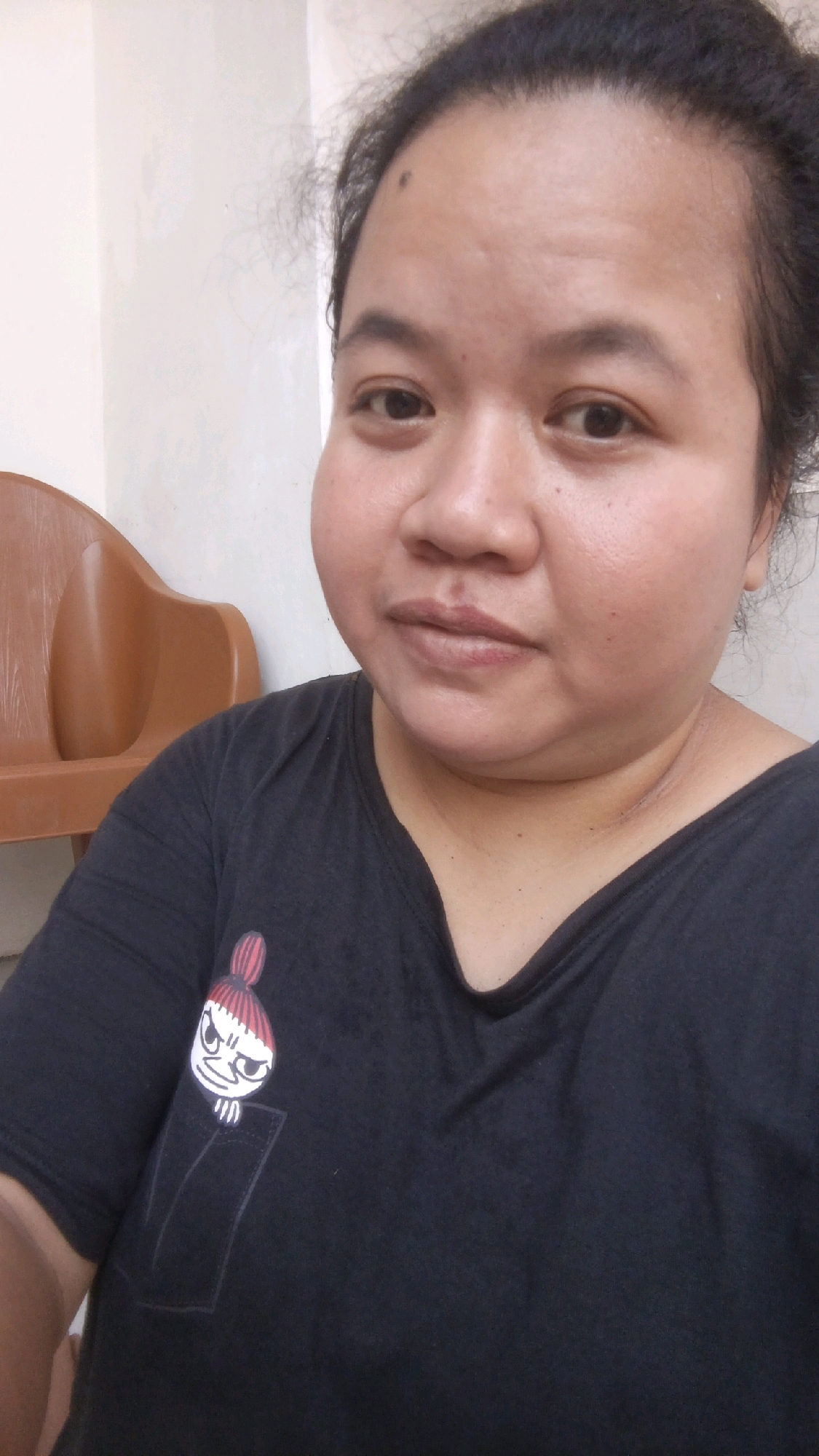 A close-up, waist-up selfie shows a person with dark hair pulled back, looking directly at the camera. The person is wearing a black t-shirt with a cartoon character peeking out of a pocket on the left side of the chest. The background is a plain, light-colored wall. To the left of the person, a brown chair is partially visible. The lighting suggests an indoor setting, likely a home or a room in Pekauman Kulon, Indonesia.