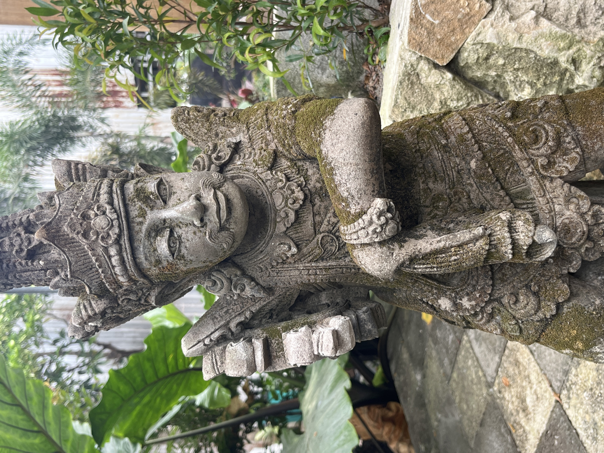 A stone statue, covered in moss, stands in a courtyard in Kuta Selatan, Indonesia. The statue depicts a figure wearing a headdress and ornate clothing, with its left arm bent and holding a draped object. The statue is positioned in front of a backdrop of lush green foliage, including large-leafed plants and trees, and a textured wall made of natural stone. The ground is paved with square stones. The overall impression is one of an outdoor garden setting.