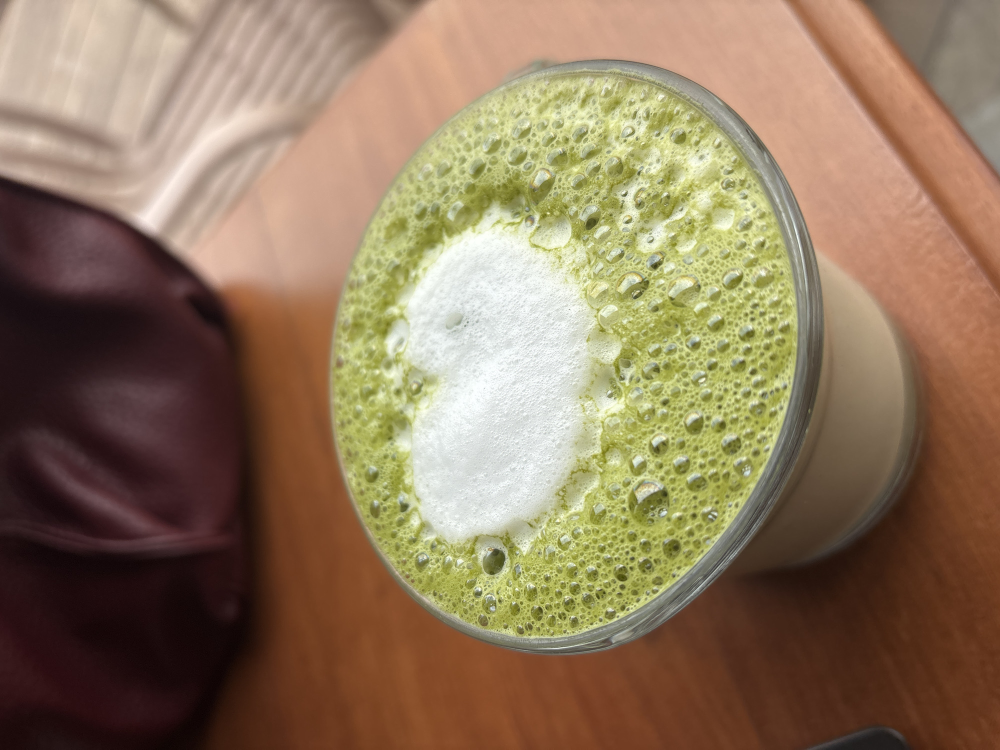 A high-angle shot reveals a glass of matcha latte on a wooden table, adorned with a heart-shaped foam design. Beside it lies a burgundy bag, partially visible in the left of the frame. The image was likely taken indoors. The city of Varna, Bulgaria, is not discernible from the image alone.