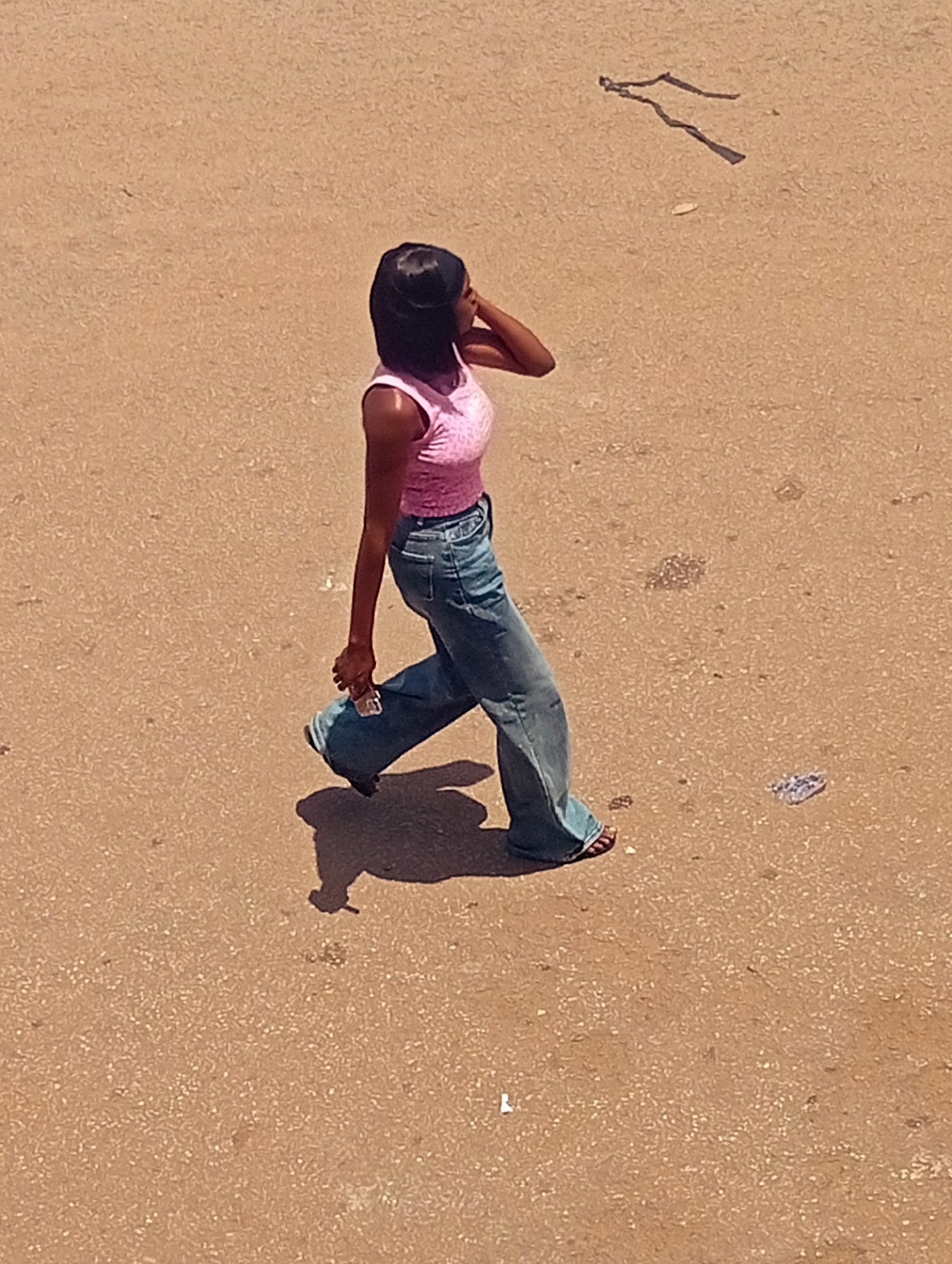 A high-angle shot shows a young Black woman walking on a tan, rough surface with minor debris scattered around. She is wearing a pink top and wide-leg blue jeans with sandals. In her right hand, she holds a small, unidentifiable object. Her left hand is raised to the back of her head or neck. The location is in the city of Unknown, Unknown.