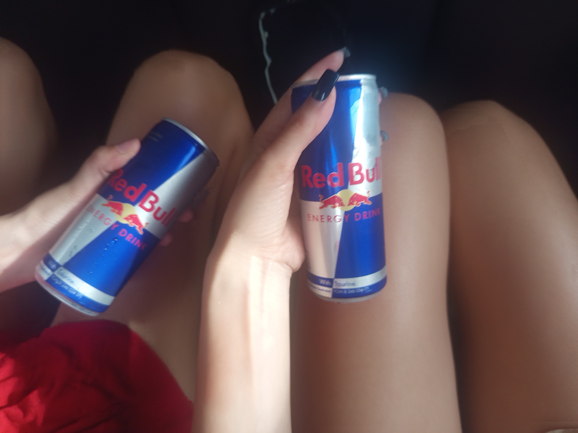 A person is holding two cans of Red Bull energy drink. The person is seated, with their thighs visible. The location is not discernible from the image.