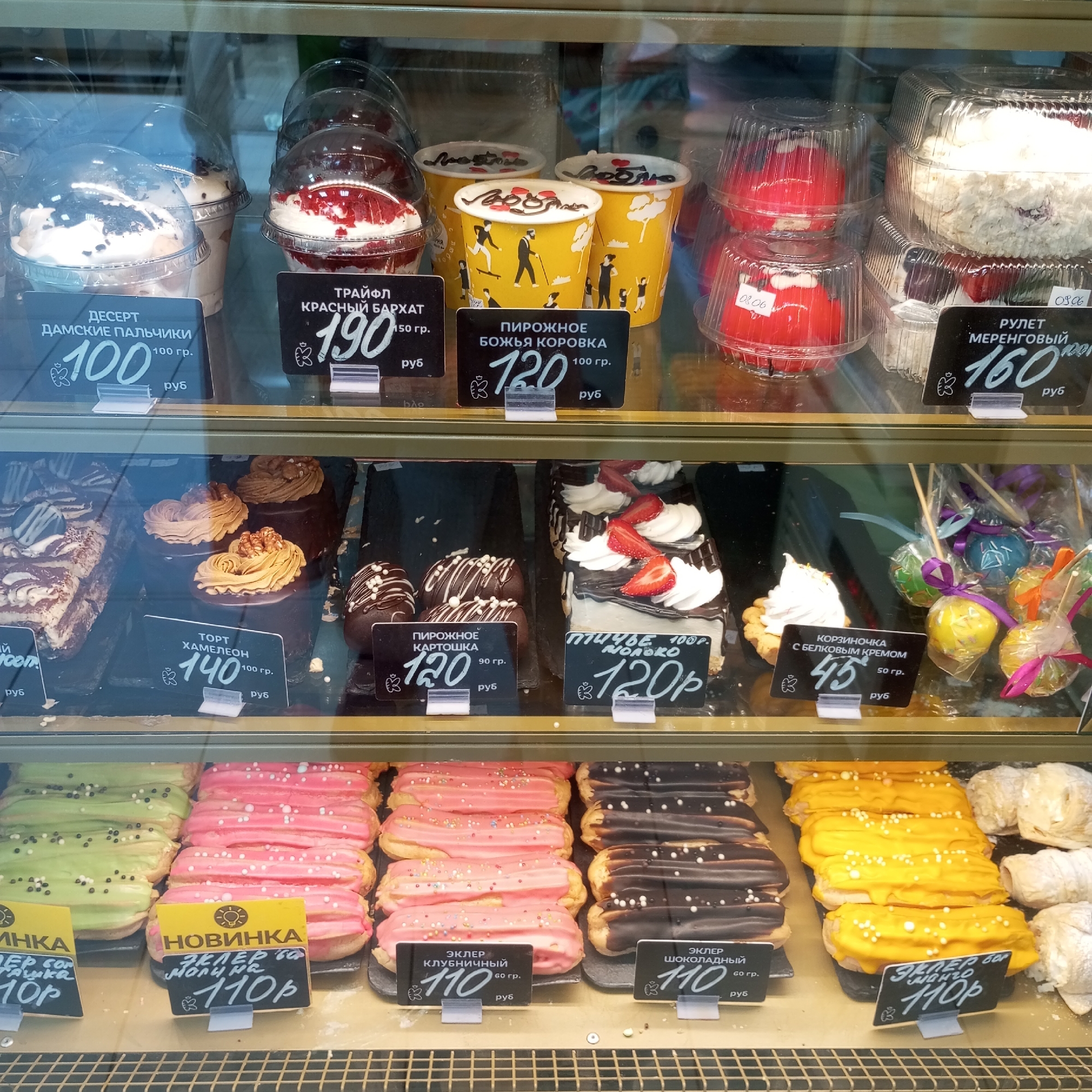 The image displays a refrigerated display case in a bakery or pastry shop in Izhevsk, Russia, filled with various desserts. The scene is viewed from the front, with the glass of the display case reflecting some of the surrounding environment.
The display case has multiple shelves. The top shelf features individual desserts in clear plastic containers, including a red velvet trifle, a dessert with chocolate shavings and cream, and yellow paper cups decorated with cartoon figures. There are also some red, heart-shaped pastries in plastic packaging and a large, white cake that appears to be a meringue roll.
The middle shelf showcases larger pastries and cakes. On the left, there is a rectangular cake with chocolate and cream layers. To its right are several dark brown, individual cakes topped with swirled frosting and walnuts. Further right, there are chocolate-covered treats with white drizzle and sprinkles, and a tart with meringue. A selection of colorful cake pops on sticks is also visible on this shelf.
The bottom shelf is filled with rows of eclairs. There are eclairs with green, pink, and dark chocolate icing, as well as yellow-frosted eclairs. Some of these eclairs have white sprinkles.
Price tags are visible in front of many of the items, indicating prices in Russian rubles (руб). Examples of prices include 100, 190, 120, 160, 140, 45, and 110. Some tags have titles such as "ДЕСЕРТ ДАМСКИЕ ПАЛЬЧИКИ" (Dessert Ladies' Fingers), "ТРАЙФЛ КРАСНЫЙ БАРХАТ" (Trifle Red Velvet), "ПИРОЖНОЕ БОЖЬЯ КОРОВКА" (Pastry Ladybug), "РУЛЕТ МЕРЕНГОВЫЙ" (Meringue Roll), "ТОРТ ХАМЕЛЕОН" (Chameleon Cake), "ПИРОЖНОЕ КАРТОШКА" (Potato Pastry), "КОРЗИНОЧКА С БЕЛКОВЫМ КРЕМОМ" (Basket with Protein Cream), and "ЭКЛЕР КЛУБНИЧНЫЙ" (Strawberry Eclair), "ЭКЛЕР ШОКОЛАДНЫЙ" (Chocolate Eclair), and "ЭКЛЕР С БОРО" (Eclair with Boron, likely a flavor).
The environment appears clean and well-lit, typical of a retail food establishment. There are no people visible in the frame. The lighting creates reflections on the glass, obscuring some details. The overall impression is a well-stocked display of sweet treats.
