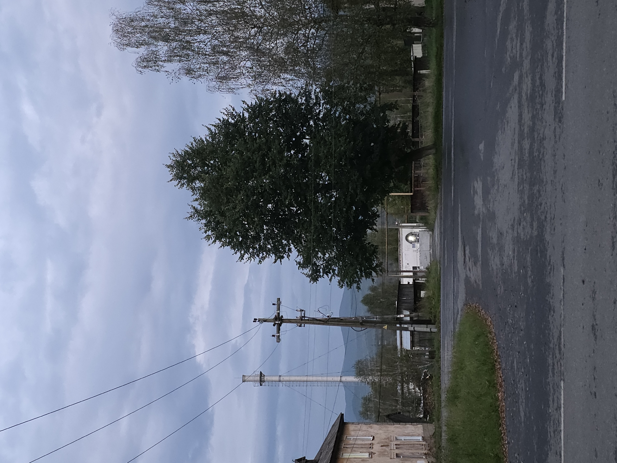 This is a vertical, eye-level, medium-shot photograph taken in Magnitka, Russia, on a cloudy day at what appears to be dusk. The image depicts a semi-rural intersection with a road curving from the lower left to the lower right.
In the foreground, the asphalt road is wet, with visible white lane markings. To the left of the road, a patch of grass with fallen leaves is visible.
Dominating the mid-ground is a large, leafy green tree. To its left, a pale, multi-story building with several windows can be seen. Further left and in the background, a tall, industrial smokestack stands against the sky. A wooden utility pole with crossbars and electrical wires is positioned between the building and the tree.
Beyond the tree, the road continues towards a gate-like structure with an arched opening, dimly lit from within. The background consists of more trees, some wooden structures, and a range of distant, hazy mountains under a cloudy sky. The overall atmosphere is subdued, with low light conditions. There are no people or animals visible in the image.