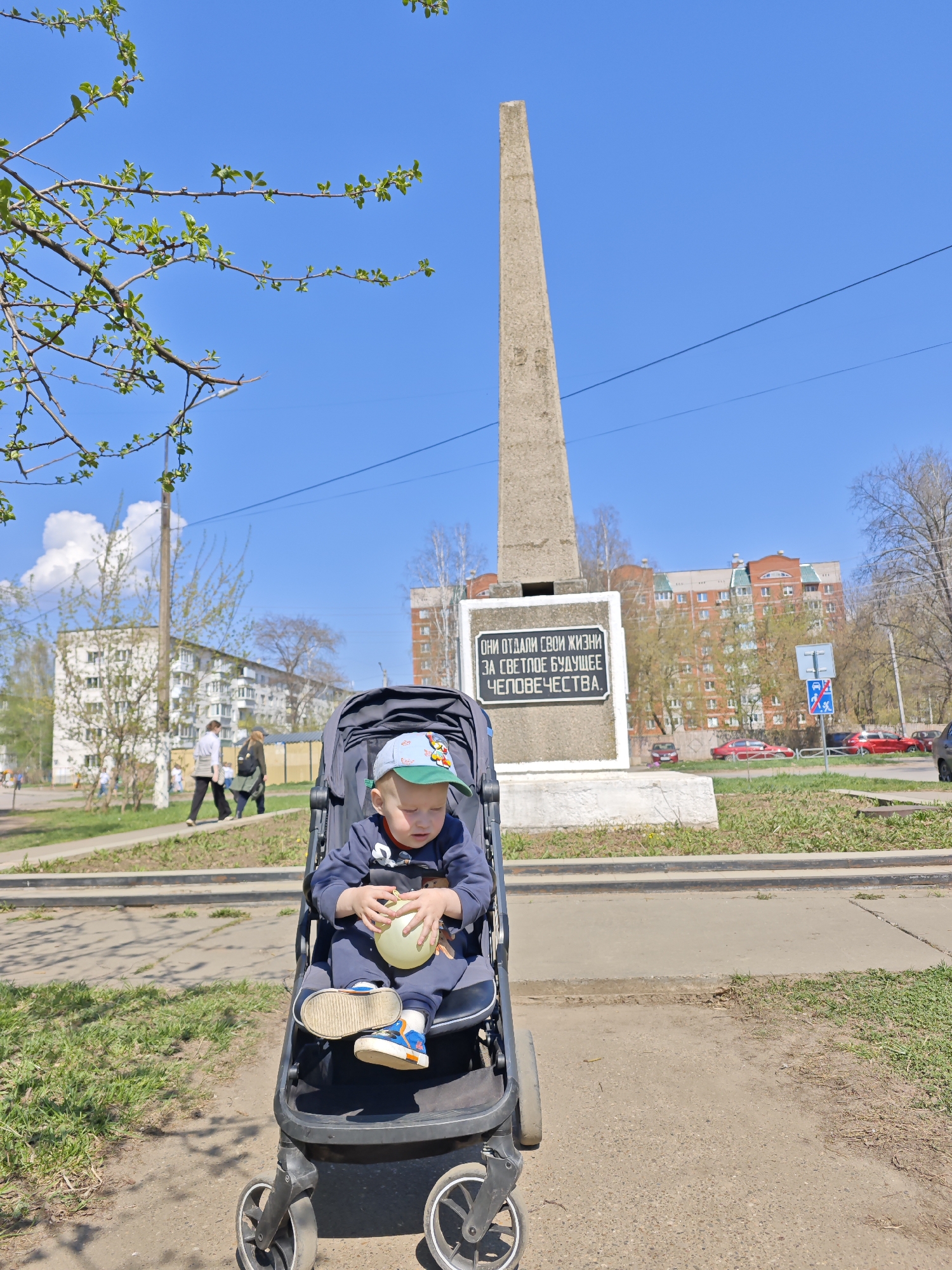 A young child sits in a black stroller on a sidewalk. The child wears a blue outfit, a green and blue hat, and blue and white shoes. The child holds a yellow balloon. Behind the child is a monument with a tall, thin, rectangular structure atop a white base. A black plaque on the base contains text. The location is Chaikovsky, Russia. In the background, a blue sky, buildings, trees, a crosswalk, and passing pedestrians are visible. Several parked red cars are near a building.