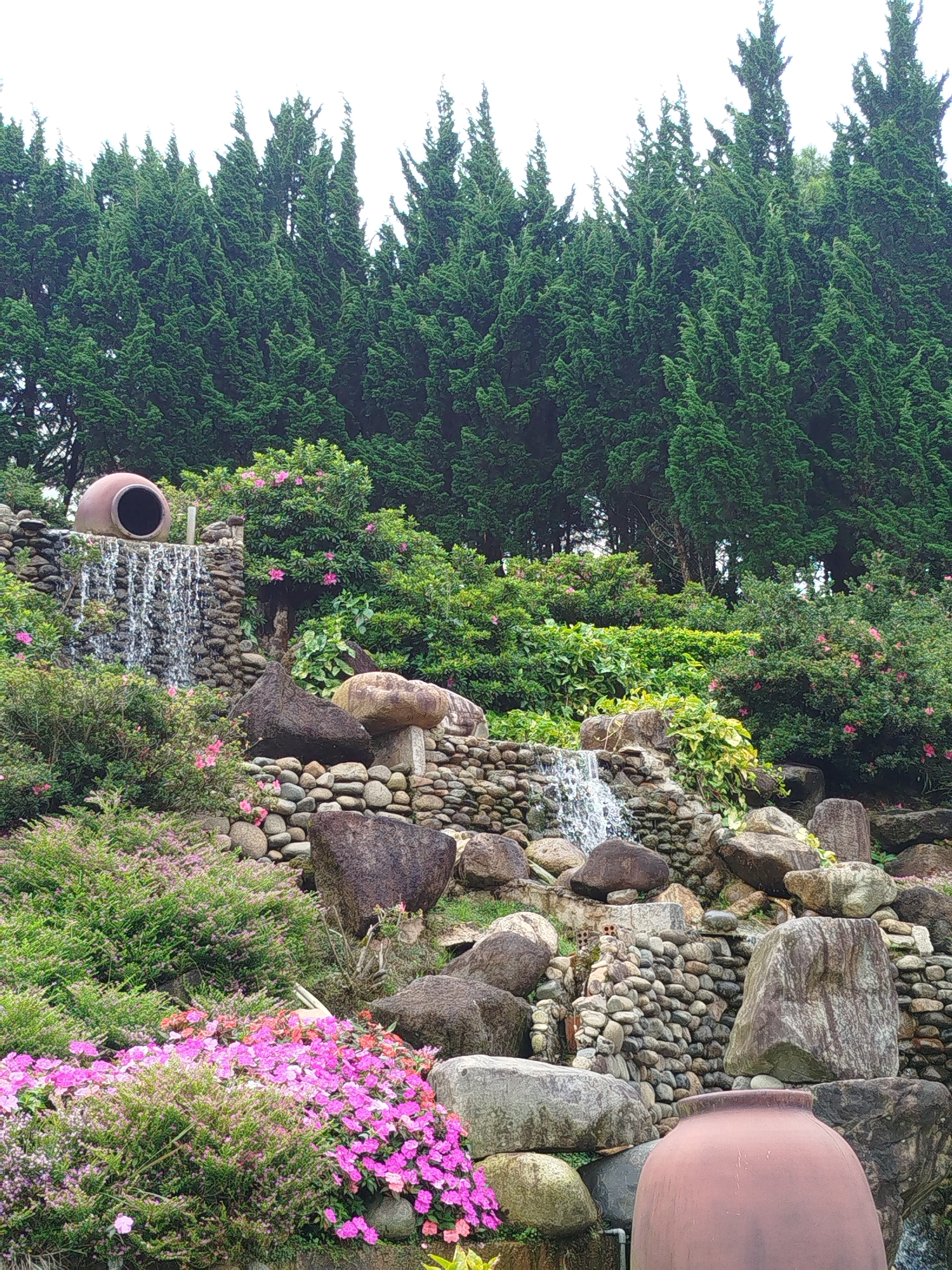 A tiered rock garden with a waterfall flows down a slope. At the top of the waterfall, a large, reddish-brown earthenware pot is partially visible, with water cascading over its rim. The garden is surrounded by lush green trees, and smaller pink and purple flowers bloom along the rocky terrain. A large, similar earthenware pot is positioned at the bottom right of the frame, partially obscuring the view. The scene is located in Lâm Viên - Đà Lạt, Vietnam.