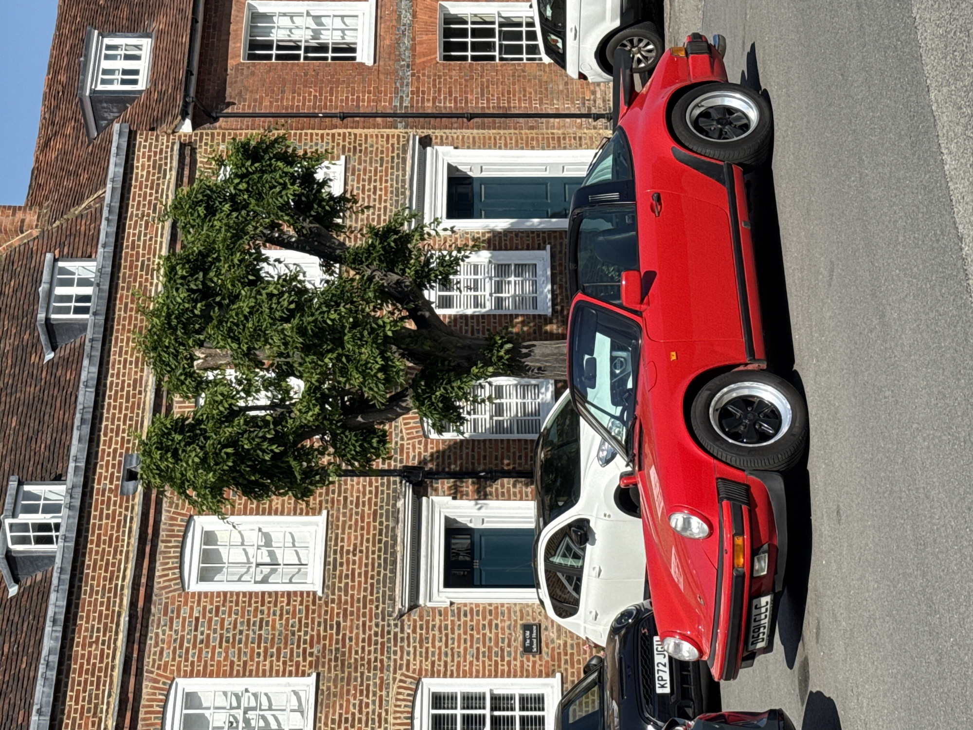 The image shows a row of parked cars on a street in Amersham, United Kingdom. The most prominent car is a red Porsche 911 parked along the sidewalk. Next to it are other parked cars, including a white Mini Cooper and a dark-colored vehicle. In the background, there is a three-story brick building with white-framed windows and a green-painted front door. A tree is visible in front of the building. The street appears to be paved with asphalt. A street sign indicating the name of the building is visible. The building is "The Old School House".