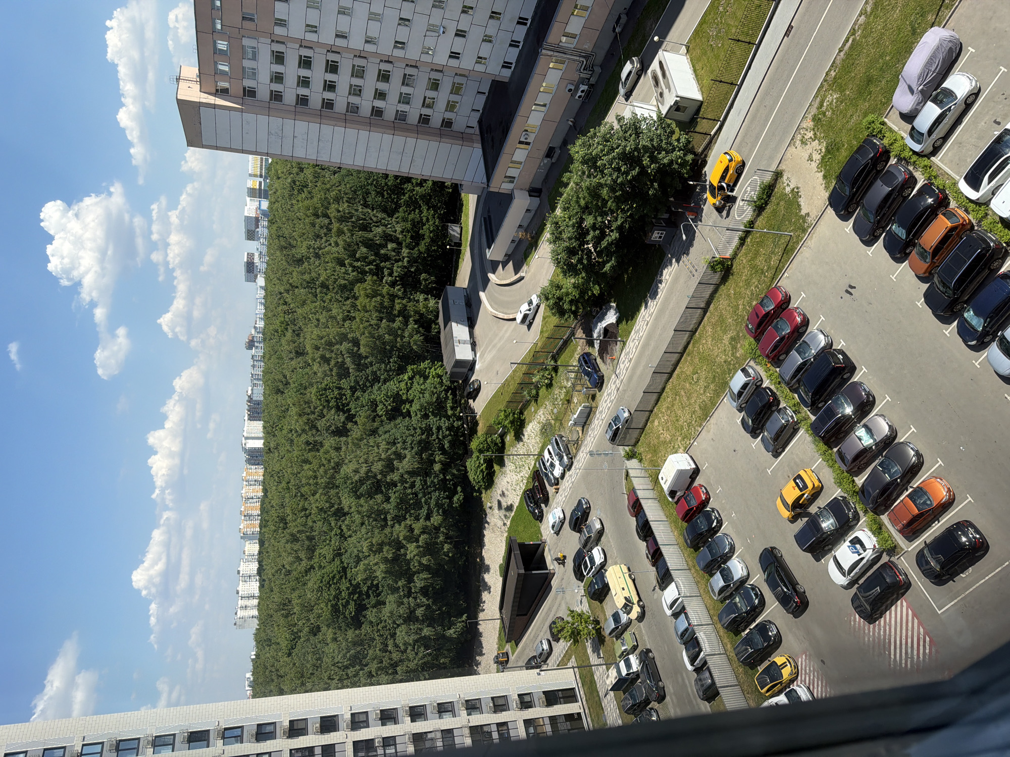 The image shows a view of a city in Moscow, Russia. The scene is a residential area with several multi-story apartment buildings, a dense forest, and a parking lot filled with various cars. The sky is blue with scattered white clouds.
In the foreground, a tall, modern apartment building dominates the left side of the frame. To the right, another apartment building, also tall and multi-story, features a distinctive pink and beige facade with many windows. Between the buildings, a parking lot is visible, containing numerous cars of different models and colors. Some cars are parked in marked spots, while others are scattered more haphazardly. A white van and a yellow vehicle are also present in the parking area. A person can be seen near the yellow vehicle, possibly interacting with it.
Behind the parking lot and buildings, a large expanse of lush green forest stretches across the middle ground. The trees are dense and appear to be a mix of deciduous species. Beyond the forest, in the background, a row of lower-rise apartment buildings can be seen, suggesting a sprawling urban landscape. The overall environment is characteristic of a suburban or urban area with significant green spaces.