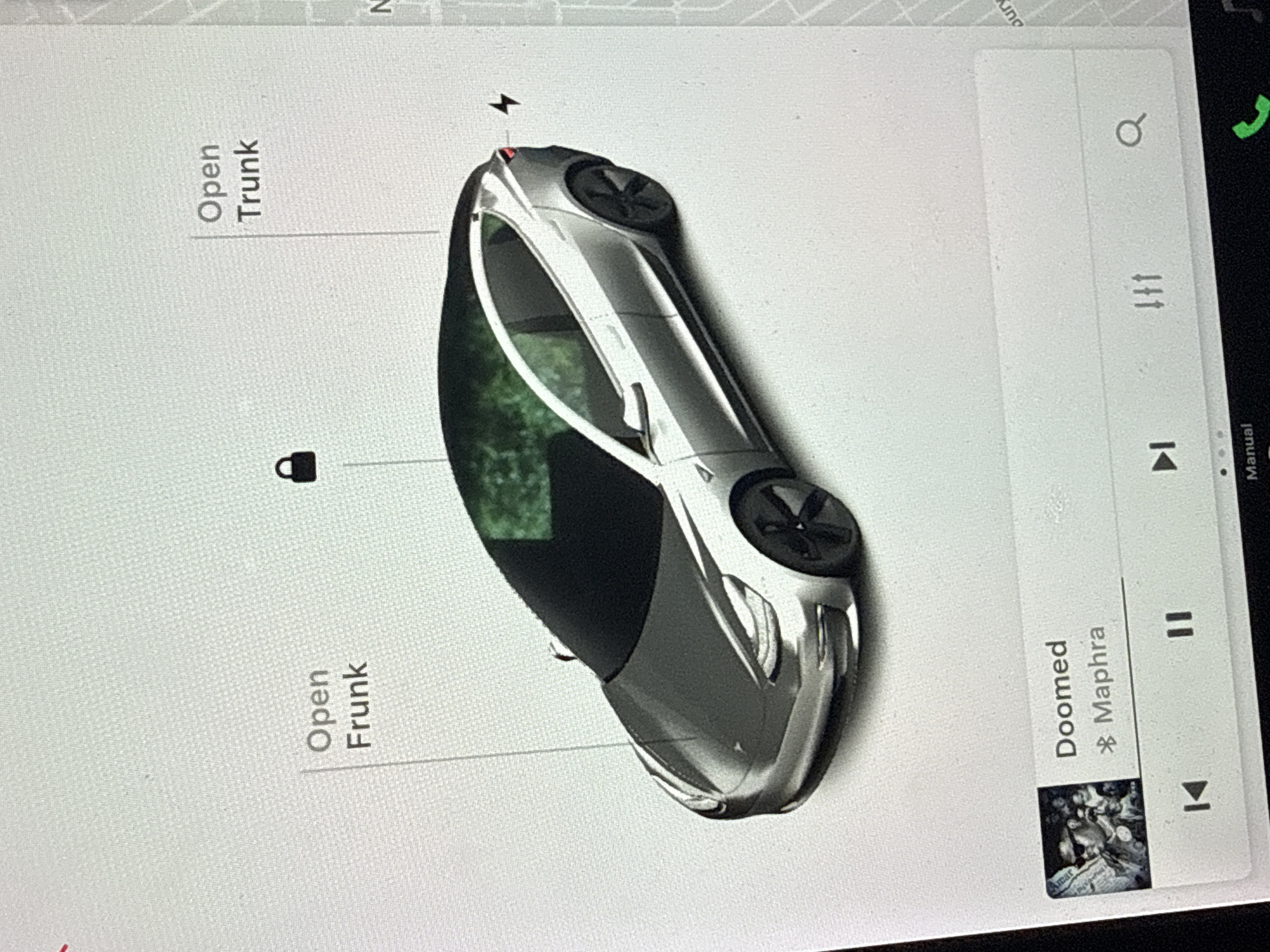 The image shows the central touchscreen display of a Tesla vehicle. The screen displays a top-down representation of a Tesla car. On the left side of the screen, there are options to "Open Trunk," and "Open Frunk." There is a padlock icon between these options. At the top of the Tesla car, there is an icon of a lightning bolt. On the right side of the screen, there is a music player showing the song "Doomed" by Mrapha playing. The screen also includes icons for search, climate control, skip, play/pause, and a phone call icon. The street on the map display is labeled “N” and “ung”, but the location is unclear. There is no evidence to locate the car in North Vancouver, Canada.