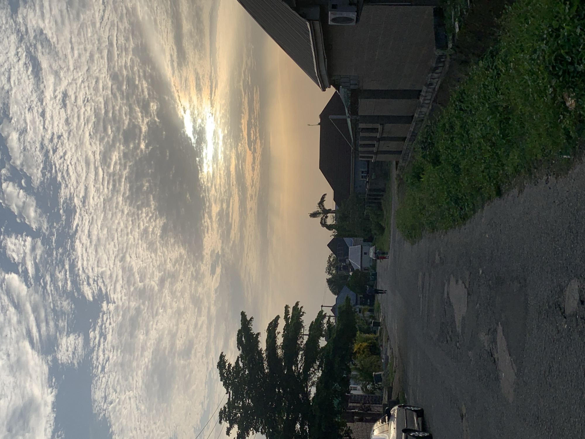 The image depicts a street scene in Abuja, Nigeria, under a partly cloudy sky with the sun obscured by clouds, creating a diffused light. The scene has a residential ambiance, featuring a paved road with signs of wear. A building is visible on the right, showing multiple floors and a possible air conditioning unit on the exterior. Lush green foliage borders the road on the right side. The street is flanked by trees on the left. In the distance, there are houses and structures along the street. A dark-colored car is parked on the left. There are silhouettes of people in the distance on the road.