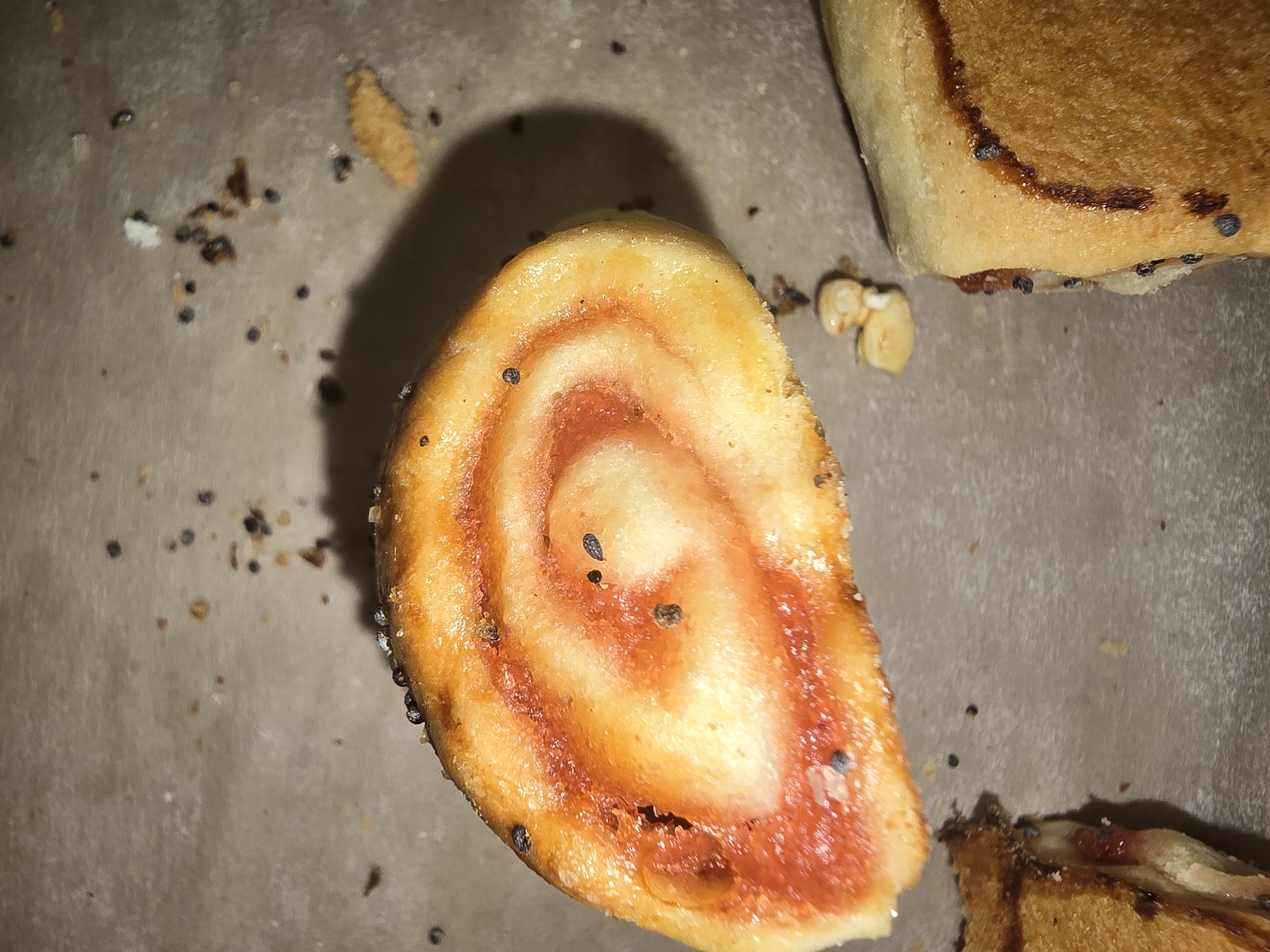 A close-up, top-down shot shows a piece of swirled pastry topped with dark seeds, likely poppy or sesame seeds, and a reddish sauce. The pastry is golden brown with a slightly uneven texture. Several of the dark seeds are visible on the surface of the pastry, with some embedded in the crust. In the background, the surface appears to be a light brown, textured material, possibly parchment paper or a similar baking surface. Scattered across this surface are more dark seeds, along with some small crumbs and other debris, suggesting it is a food preparation area. To the lower right of the frame, another piece of baked good is partially visible, appearing to be a sandwich or roll with dark seeds along its edges. The lighting is somewhat dim, casting a subtle shadow from the pastry onto the background. The image is focused on the pastry and its immediate surroundings, providing a detailed view of the food item and the surface it rests on.
This image is a close-up of food items and is not located in Ulan-Ude, Russia.