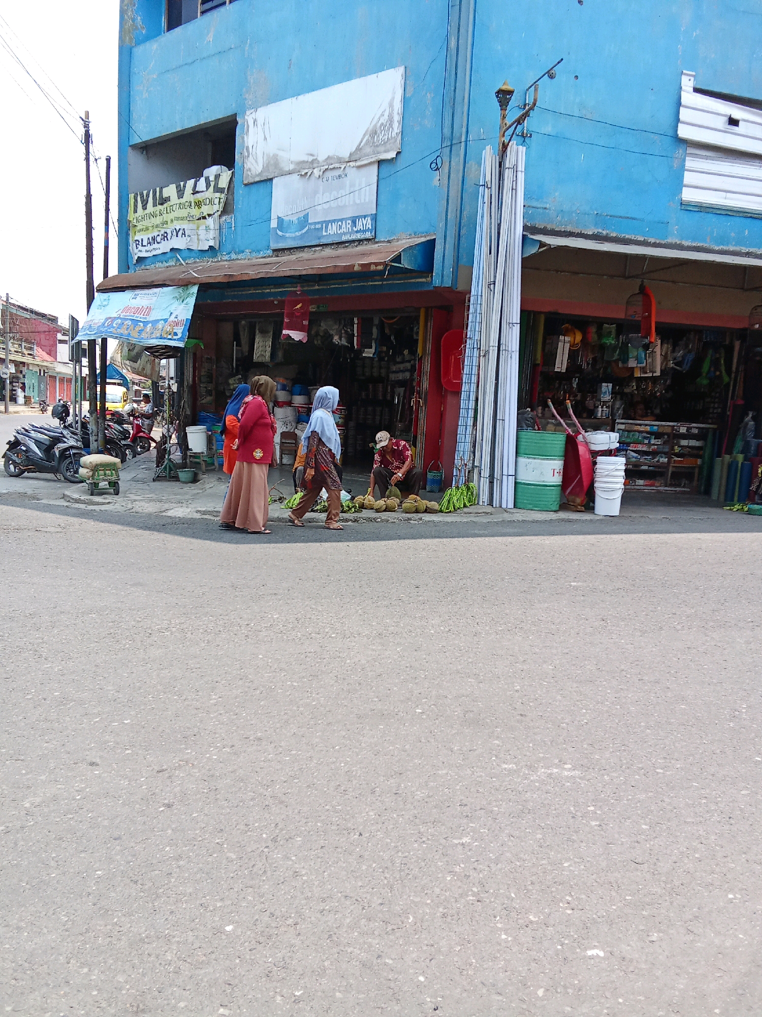 This is a ground-level, medium shot of a street scene in Banjarnegara, Indonesia, on a bright, overcast day. A blue, two-story building with a corrugated metal roof juts out into the frame from the right, housing several storefronts. One storefront, labeled "IVIL VSIL LIGHTING & ELECTRICAL PRODUCT" and "BLANCAR JAYA", is partially obscured by awnings and displays. Another storefront, labeled "LANCAR JAYA", features a blue shoe advertisement.
In the foreground, a paved street occupies the lower half of the image. To the left, several motorcycles are parked along the sidewalk. A small, green, wheeled cart sits near the motorcycles.
On the sidewalk in front of the building, three individuals are visible. Two women, one in a red top and beige hijab and the other in a blue hijab and patterned scarf, walk past a man sitting on the ground. The man, wearing a red and white striped shirt and a white cap, is arranging a pile of durian fruit. A third person, partially obscured, walks away from the viewer in the distance.
Various objects are visible on the sidewalk and in front of the shops, including stacks of white PVC pipes, green barrels, white buckets, gardening tools, and bags of what appear to be cement or fertilizer. The overall impression is of a bustling commercial street.