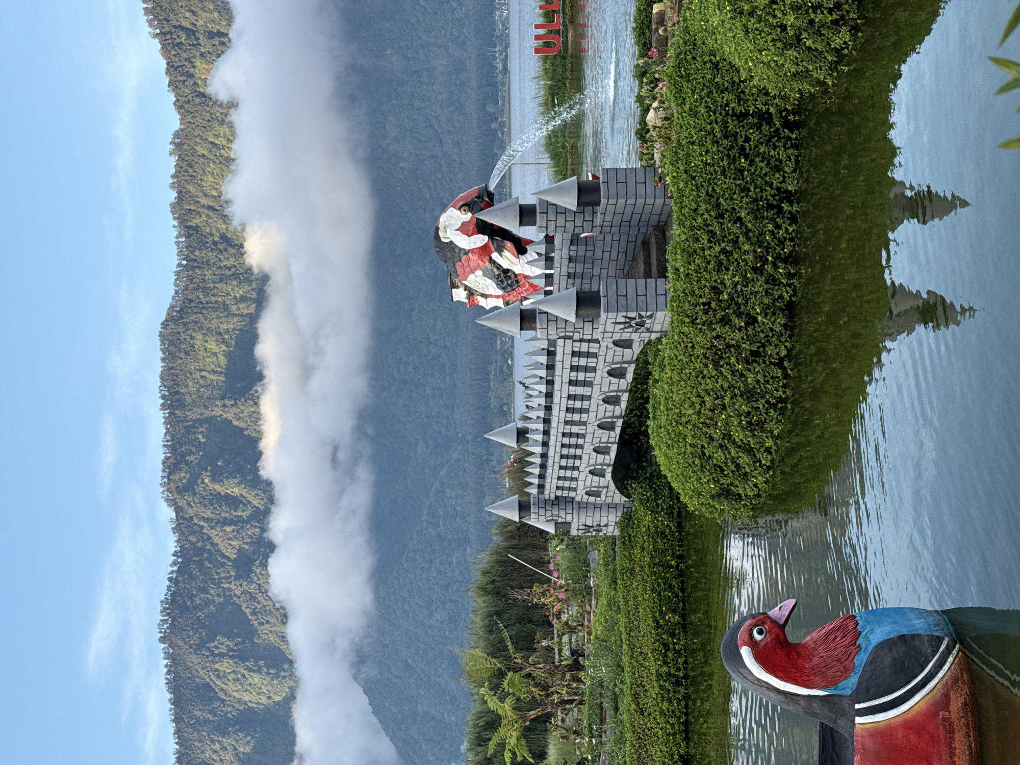 The image shows a park scene in Baturiti, Indonesia, featuring a whimsical architectural display in a body of water. A castle-like structure with grey stone-patterned walls and white pointed turrets is topped by a large fish-shaped sculpture. The fish is colored with red, white, and black patterns, and water spouts from its mouth. The architectural display is surrounded by carefully manicured hedges and vegetation. A colorful duck statue sits in the foreground near the edge of the water. The background features a landscape of mountains partially obscured by clouds. The image appears to be taken during daylight hours.