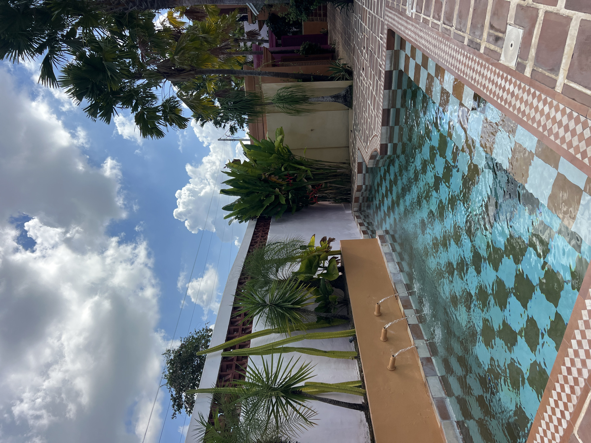 The image depicts an outdoor courtyard with a swimming pool in North Kuta, Indonesia. The pool features a checkered tile pattern in shades of blue and brown. Three terracotta spouts are visible on an orange wall, from which water is flowing into the pool, creating ripples on the surface. Surrounding the pool is a tiled patio with a decorative border. Lush green plants, including palm trees and what appear to be tall, cylindrical cacti, are planted along the wall behind the pool. In the background, a building with colorful accents, including a pink and purple facade, is visible. The sky above is blue with scattered white clouds. No people are present in the scene.