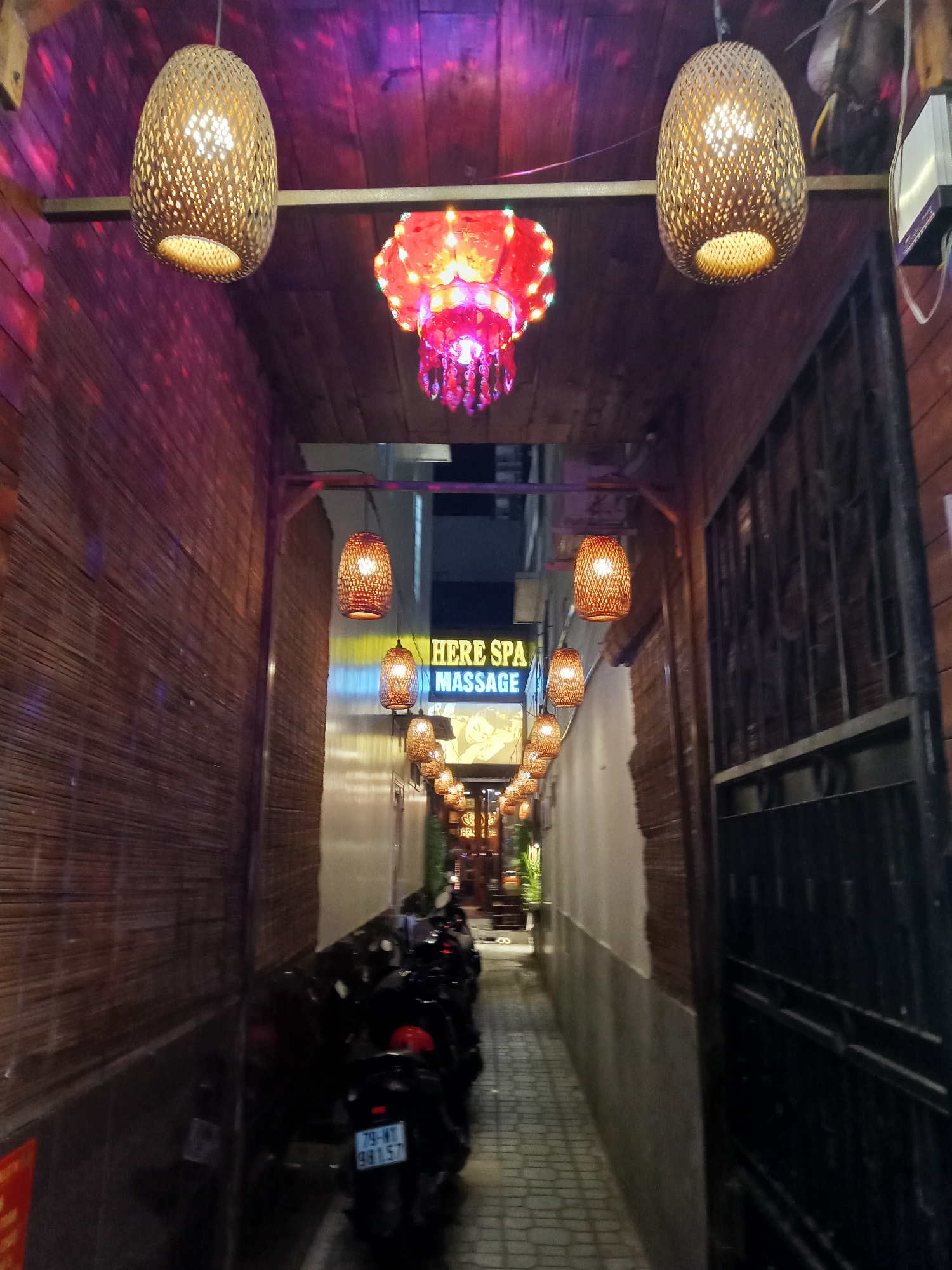 A narrow street in Nha Trang Ward, Vietnam, is illuminated by a series of hanging lights. The street is lined with buildings on either side. Parked motorbikes sit along the right side of the street. A sign for "HERE SPA MASSAGE" is visible in the distance. The lighting creates a warm and inviting atmosphere.