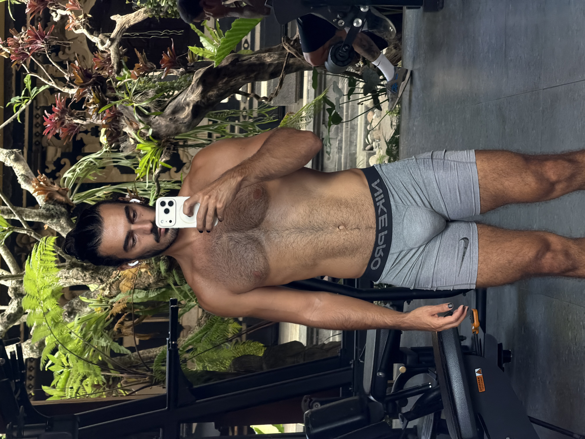 A shirtless man with dark hair pulled back in a bun, wearing gray Nike boxer briefs, stands in what appears to be an outdoor gym area within a lush, tropical setting, possibly at a resort or spa in North Kuta, Indonesia. He holds a white smartphone in his right hand, taking a selfie in a mirror. He has earbuds in his ears and dark nail polish on his fingernails. The background features dense green foliage, a wooden structure adorned with plants, and gym equipment, including a weight machine partially visible on the left. Another person is faintly visible in the background on the right, seated on gym equipment. The floor is tiled with large gray tiles.
