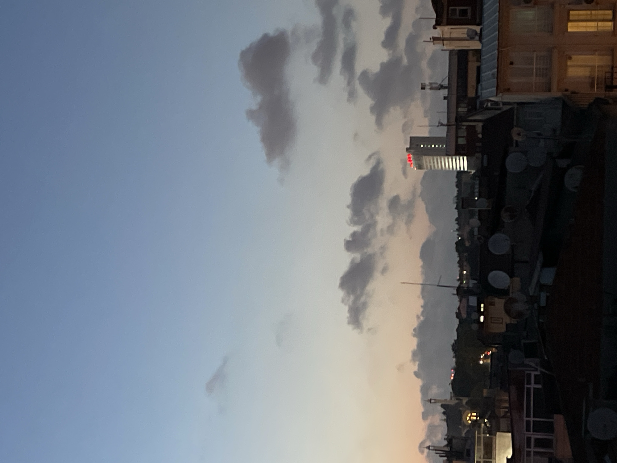 This image captures a twilight view of the city of Kadıköy, Turkey, from an elevated perspective, likely a rooftop. The sky transitions from a pale blue at the top to soft oranges and pinks near the horizon, with scattered clouds adding texture. In the background, a modern, white skyscraper stands out, illuminated by vertical white lights and topped with prominent red illuminated signage that reads "HALKBANK." Below the skyscraper, a mix of buildings, some with illuminated windows and satellite dishes, create a dense urban landscape. A slender minaret is visible amidst the lower buildings, suggesting the presence of a mosque. The foreground consists of dark rooftops and the upper floors of buildings, with some lights visible within them. The overall scene conveys a sense of urban life settling into the evening.