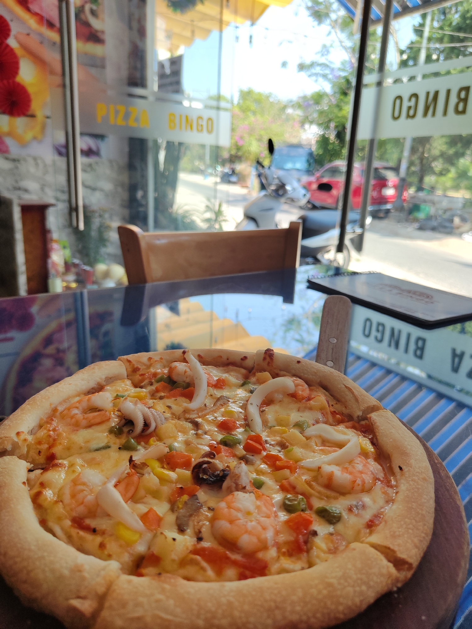 A high-angle, indoor shot captures a seafood pizza served on a wooden board at a table in Pizza Bingo in Da Lat, Vietnam. The pizza features a thick crust, melted cheese, shrimp, squid, and chopped vegetables. Behind the pizza, the restaurant's interior is visible, with a wooden chair and a glass tabletop reflecting the surroundings. Through the glass windows, a street scene unfolds, including a scooter and red car. A black menu sits at the side of the table. The glass door displays the words "Pizza Bingo."