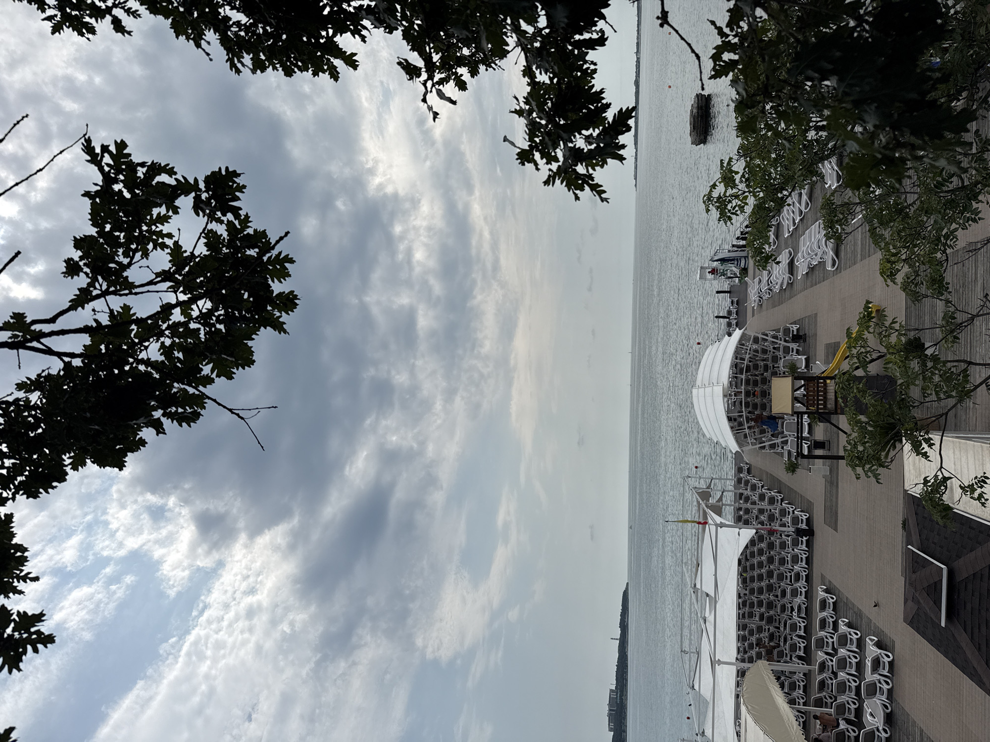 The image captures a view of a beach resort in Gelendzhik, Russia, on an overcast day. The foreground is partially obscured by the dark, leafy branches of trees, creating a natural framing effect. Below the trees, a wooden boardwalk stretches out towards the sea, lined with rows of white lounge chairs neatly arranged under white canopies. These canopies provide shade for the seating area.
Further down the boardwalk, there is a covered structure that appears to be a cafe or restaurant, with some patrons visible sitting at tables. A small yellow slide is set up near this structure, suggesting a family-friendly environment. Beyond the boardwalk and seating area is the calm, gray expanse of the sea, meeting a hazy horizon. In the distance, a town or settlement with buildings and a lighthouse can be seen on the left side of the image. The sky is filled with a mix of white and dark gray clouds, indicating overcast weather. The overall scene depicts a seaside resort preparing for or winding down from a day of activity under a cloudy sky.