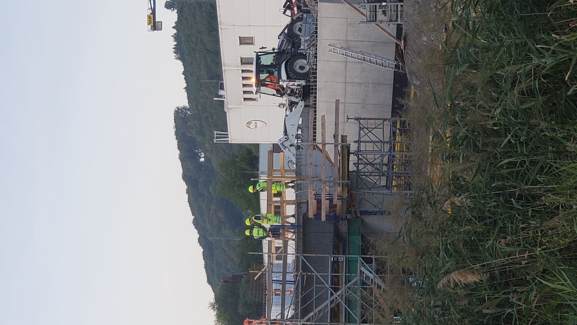 The image depicts an outdoor construction site, likely on a slightly overcast day. The foreground is dominated by tall, green reeds and grasses. Behind the vegetation, a concrete bridge structure is under construction, supported by scaffolding. Several construction workers, wearing high-visibility yellow vests and helmets, are present on the scaffolding. One worker is standing, seemingly observing. Two others are standing closer together, possibly conferring.
On the bridge deck, a white backhoe loader is parked. A construction worker in an orange vest is visible inside the cab of the loader. To the right of the loader, a ladder is leaned against the bridge structure, and further to the right, there are mounds of dirt and construction materials.
In the background, a modern, white building with multiple windows and a circular logo is visible. The building appears to be a commercial or industrial facility. Behind the building, a tree-covered hillside rises, and a blue crane arm with a yellow "WART" logo is partially visible against the pale sky. The overall scene suggests an active construction project in an area with a mix of industrial and natural elements. The specific location in the city of Prešov, Slovakia, cannot be determined from the image alone, as there are no identifying landmarks or text present in the scene that specifically pinpoint its location within Prešov.