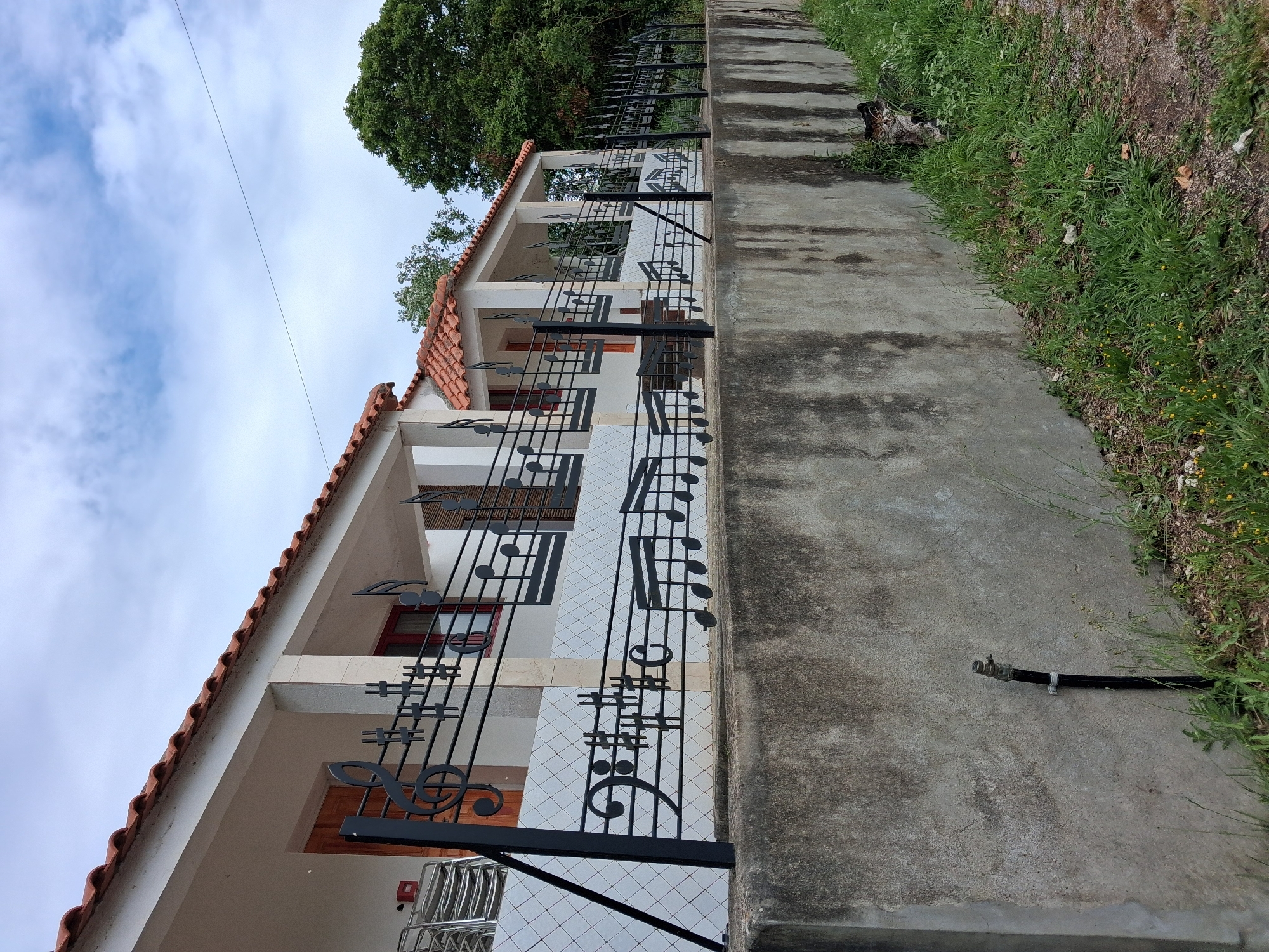 A low-angle, color photograph depicts the exterior of a two-story building in Soure, Portugal. The building features a white façade with a red-tiled roof. The railing is decorated with black ironwork musical notes and staff lines. The building has multiple evenly spaced windows with visible red frames. The building sits atop a gray concrete wall. Beyond the wall, a strip of grass separates the wall from the ground. The sky is overcast with a few patches of blue visible. A tree is visible above the building. Power lines are seen near the left edge.