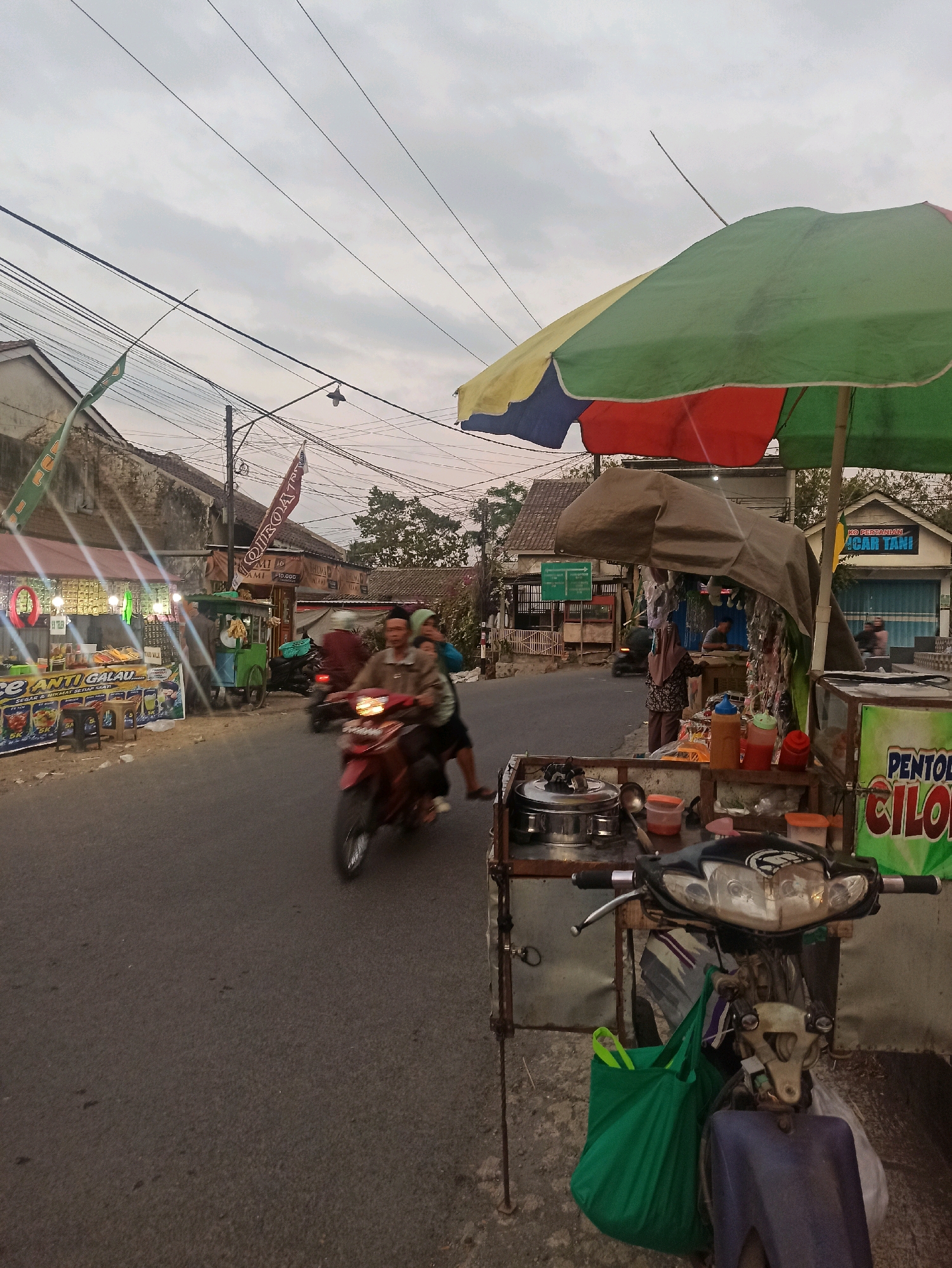 This is a street-level, medium-shot, eye-level, slightly angled, natural-light photograph taken at dusk in Tlogosari, Indonesia. The scene depicts a street with various vendors and activity. In the foreground, a food cart is parked, with a scooter in front of it. The food cart is covered by a multicolored umbrella and has various containers and food items visible. A green shopping bag is attached to the scooter.
The street is paved and appears to be moderately busy. A red motorcycle carrying two people is in motion, passing by the food cart. The rider is wearing a brown jacket, and the passenger is wearing a blue headscarf. On the left side of the street, there are several stalls with illuminated signs, suggesting food or merchandise being sold. One sign prominently displays "ANTI GALAU" with images of drinks. Further down the street, a green pushcart vendor is visible.
The background shows buildings and trees. Overhead, there is a complex network of power lines and wires. The sky is overcast, typical of the late afternoon or early evening. The overall impression is that of a lively street scene in a residential or commercial area of Tlogosari.