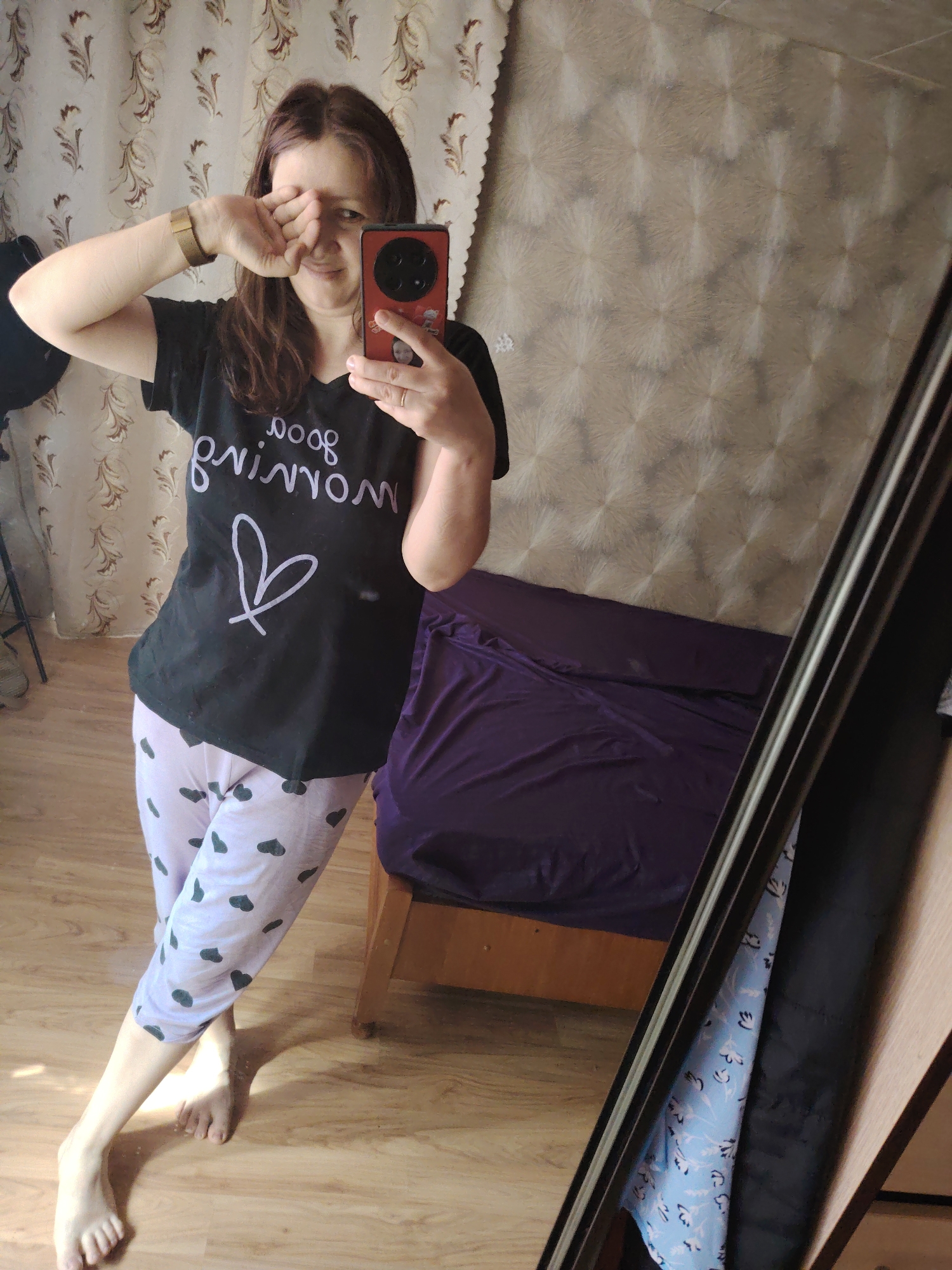 A woman is taking a mirror selfie in a bedroom. She is wearing a black t-shirt with white writing and a heart, and light purple pajama pants with black hearts. Her right arm is raised, covering part of her face. She is holding a red smartphone in her left hand, which is angled to capture the reflection. The background shows a patterned wallpaper, a bed with purple bedding, and a wooden floor. The scene appears to be indoors, likely a residential setting. The question about the city of Rostov-on-Don, Russia is not supported by any visual evidence in the image.