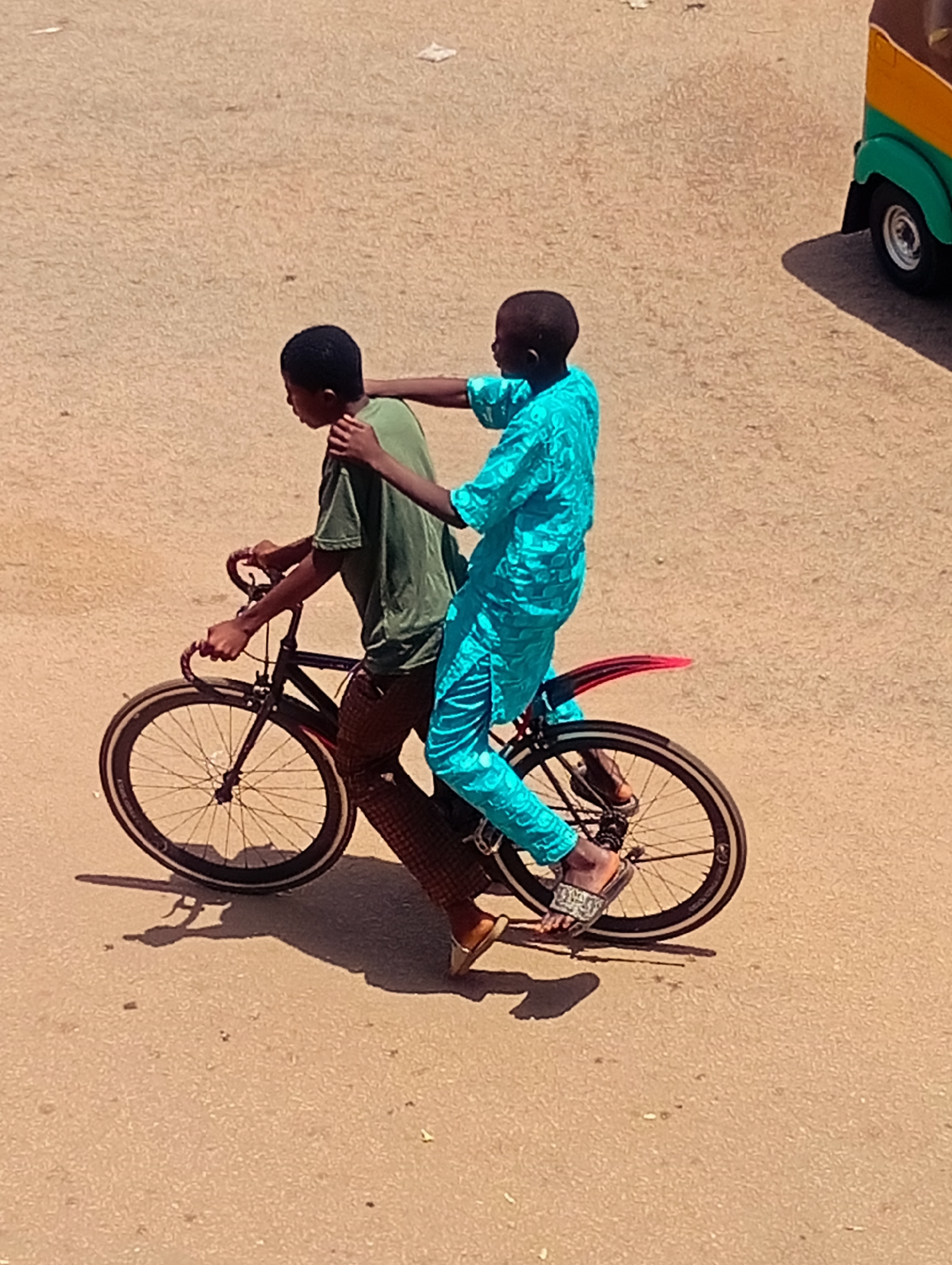 In Bukuru, Nigeria, two young boys are captured in a moment of shared transport. The boy in the front, wearing a bright turquoise kaftan, pedals a bicycle while the boy behind him, dressed in a green t-shirt and patterned brown trousers, clings to his shoulders for support. The bicycle, a simple black frame with spoked wheels, is in motion on a dusty, sun-baked street. The background is indistinct, a blur of pale earth and a hint of a colorful three-wheeled vehicle parked to the right, its shadow stretching across the ground. The scene is illuminated by bright sunlight, casting sharp shadows that emphasize the texture of the road.