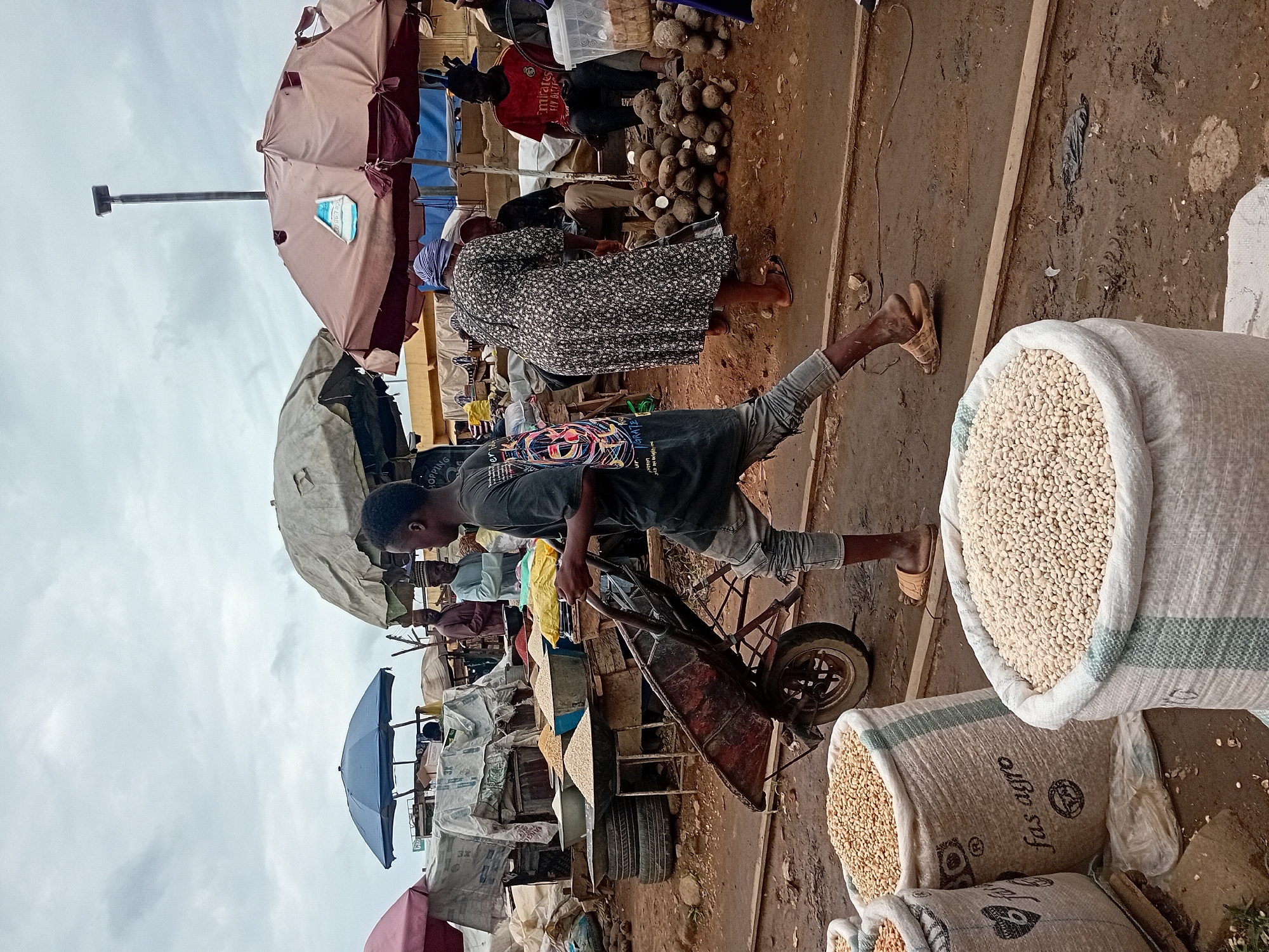 This is a medium shot, eye-level, outdoor photograph taken in a marketplace in Maiburiji, Nigeria, under a cloudy sky. In the foreground, three large, white sacks are partially filled with light-colored beans. The sack on the right is the most prominent, with green stripes. A person, identified as a young male, is walking from left to right, pushing a red wheelbarrow laden with goods. He is wearing a dark t-shirt with a colorful graphic and light-colored distressed jeans. His feet are bare except for simple sandals. Behind him, a woman in a dark, floral-patterned dress is standing, facing away from the camera, and appears to be interacting with a stall. To her left, a man in a light blue shirt and a patterned cap is also present. Further in the background, several market stalls are visible, some covered by large, beige and brown umbrellas. These stalls display various goods, including grains in large bowls, root vegetables piled on the ground, and other unidentified items. A tall, slender pole with a light fixture stands prominently against the sky. The ground of the marketplace is dirt and appears to be wet in places. The overall scene depicts a bustling, outdoor market environment.