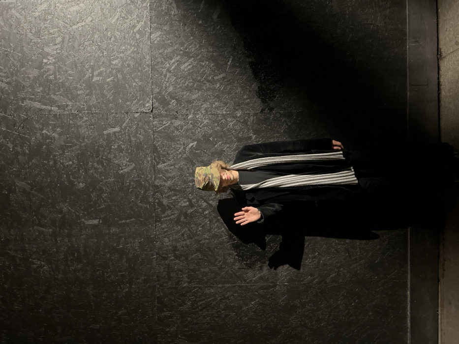 The scene is located in Paris, France, featuring a single individual standing against a dark, textured wall. The wall is composed of large, rectangular panels of what appears to be black-painted oriented strand board (OSB), with visible seams forming a grid pattern. The individual, a male with light brown wavy hair, wears a camouflage-patterned bucket hat, a dark grey t-shirt, a dark velvet long-sleeved jacket, black trousers, and a vertically striped grey and white scarf. His left arm is raised, palm open and facing forward, in an apparent greeting or stop gesture. A faint smile is present. Lighting originates from the left side of the frame, casting a distinct, elongated shadow of the individual onto the wall to his left. The lower section of the scene, including the individual's lower body and the ground, is heavily shadowed. A narrow strip of light-colored ground material, consistent with paving, is visible at the very bottom.