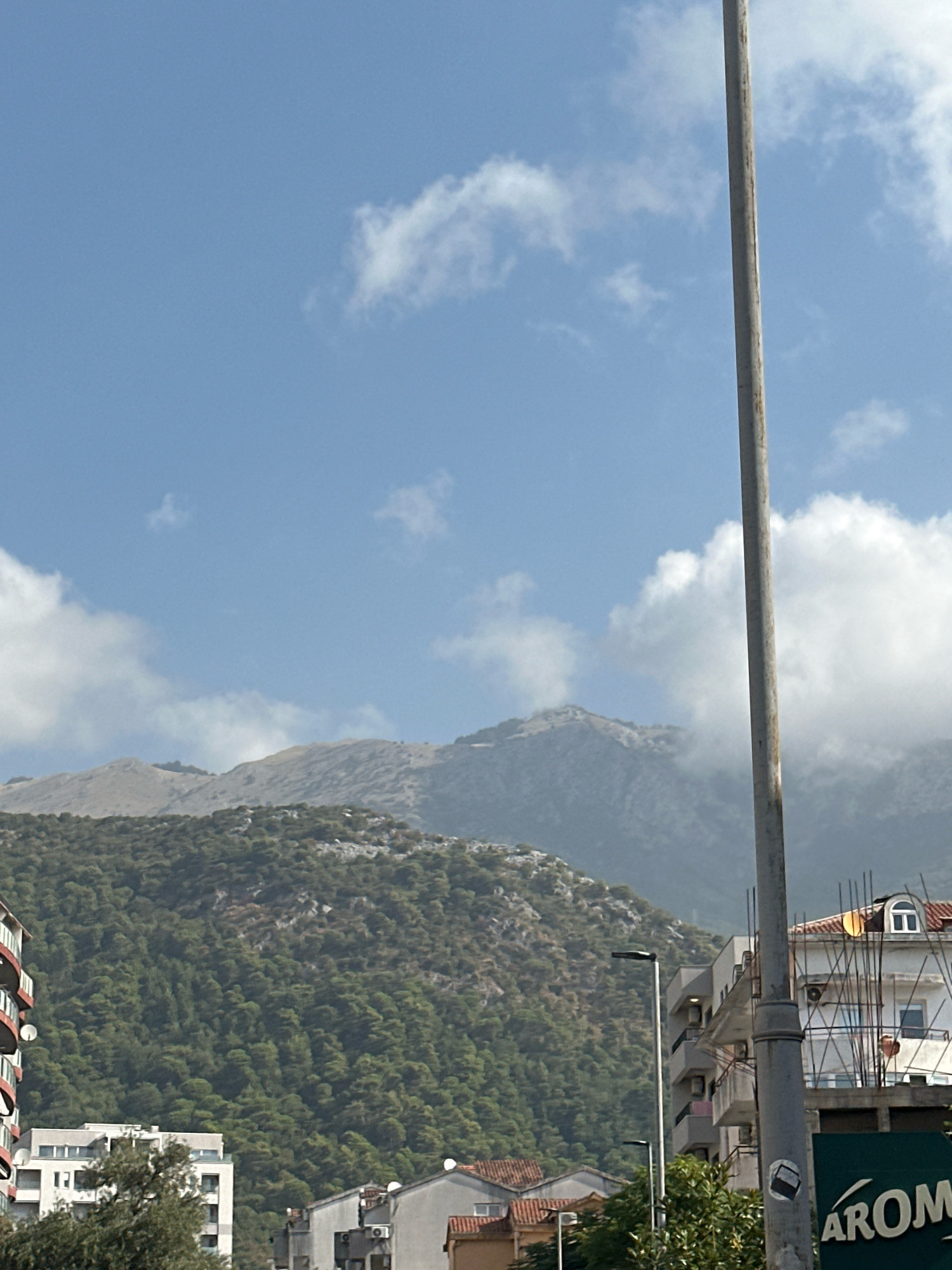 The image captures a view of the mountainous terrain behind the city of Budva, Montenegro, under a clear blue sky dotted with white clouds. In the foreground, a tall, weathered utility pole stands prominently, bisecting the frame vertically. To the left, parts of buildings are visible, including a section of a red-and-white striped structure with balconies, and a modern white building with multiple windows. In the lower right, another building with a white facade and arched windows is present, with exposed rebar suggesting ongoing construction or disrepair. Partially obscured by the pole is a green sign with white lettering that reads "AROM". The middle ground is dominated by a dense, green forested slope that transitions into a more barren, rocky mountainside further in the distance. The top of the mountains is partially covered by wisps of clouds, adding depth to the scene. The overall impression is one of urban development at the foot of a rugged natural landscape.