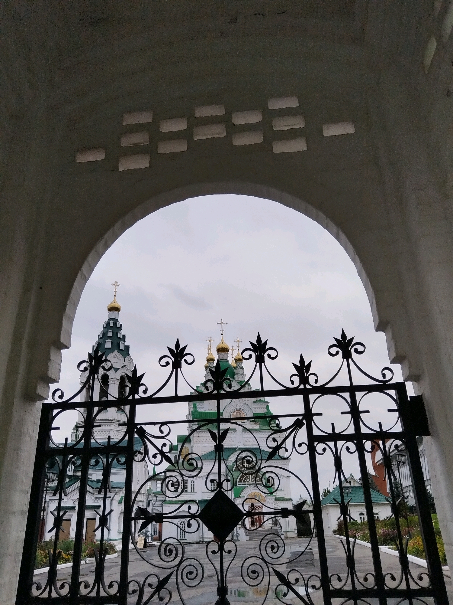 The image shows an architectural view through an arched entryway with a decorative metal gate in the foreground. The gate is black and ornate, featuring swirling metalwork and pointed finials. Beyond the gate, a white building with green roofs and golden domes is visible, likely a church or cathedral. The sky is overcast. The building appears to be the Annunciation Cathedral in Yoshkar-Ola, Russia. The arched entryway frames the scene, with a textured white surface on the left and a rough, greyish ceiling with rectangular cutouts above. The cutouts are arranged in two rows and appear to be gaps in the construction, allowing some light or air to pass through. The overall impression is one of entering a sacred or historic site.