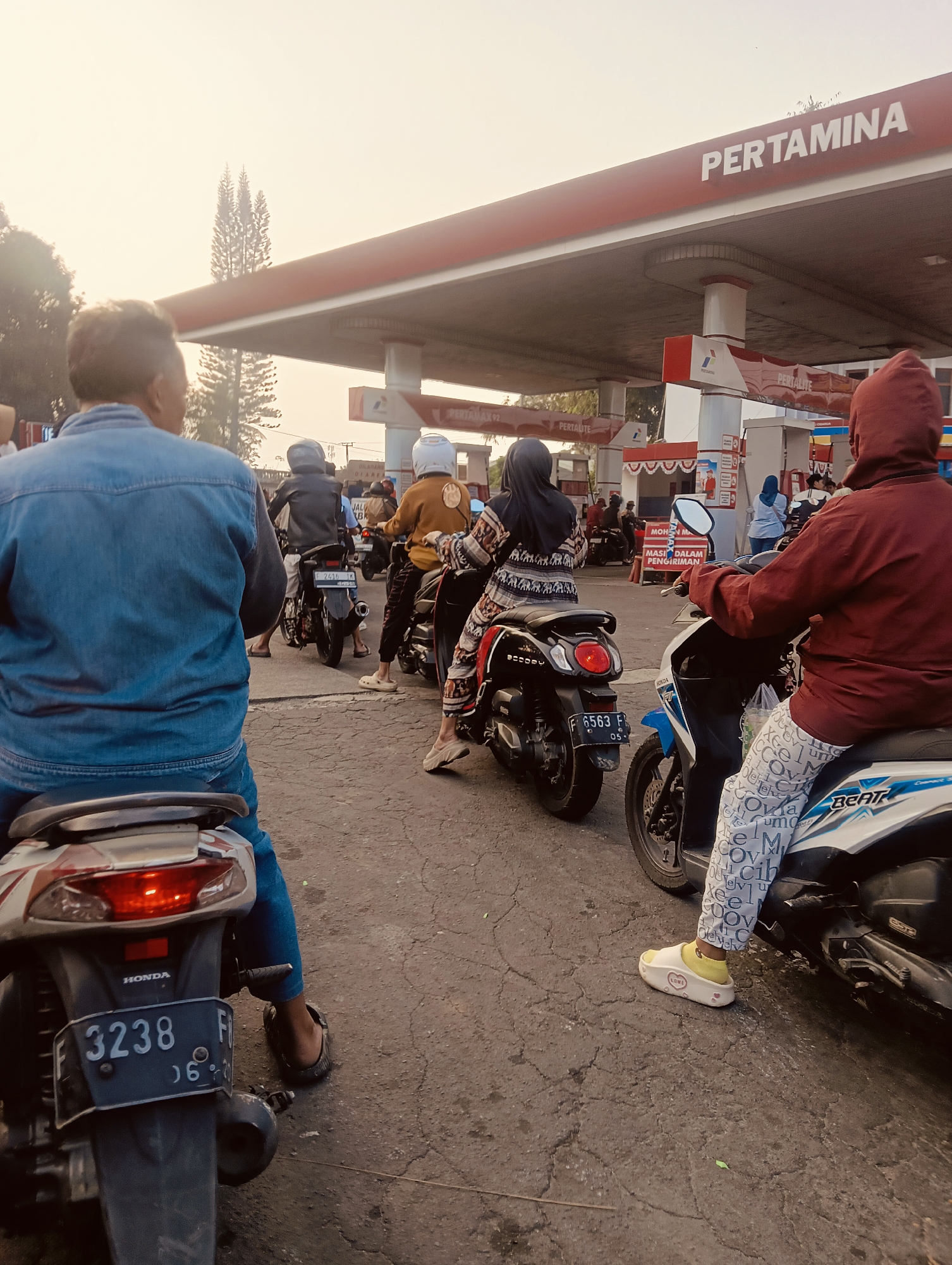 The image depicts a queue of motorbikes at a Pertamina gas station in Cisarua, Indonesia. The scene is captured from a low angle, focusing on the rear of several motorbikes and their riders. The sky is overcast with a hazy, warm light, suggesting it might be early morning or late afternoon.
In the foreground, a person wearing a denim jacket is seated on a motorbike, facing away from the camera. To their right, another person in a maroon hooded jacket rides a white and blue motorbike, wearing patterned pants and white slip-on shoes with yellow socks. Further down the queue, a person in a patterned dress and hijab is on a black scooter. Behind them, a person wearing a light brown jacket and a white helmet rides another motorbike, also with a person in a hijab behind them.
The Pertamina gas station canopy with its red and white signage is visible in the background. Gas pumps and other station equipment are also present. The ground is paved with asphalt that shows signs of wear and cracking. The overall atmosphere suggests a typical day at a busy refueling station in Indonesia.