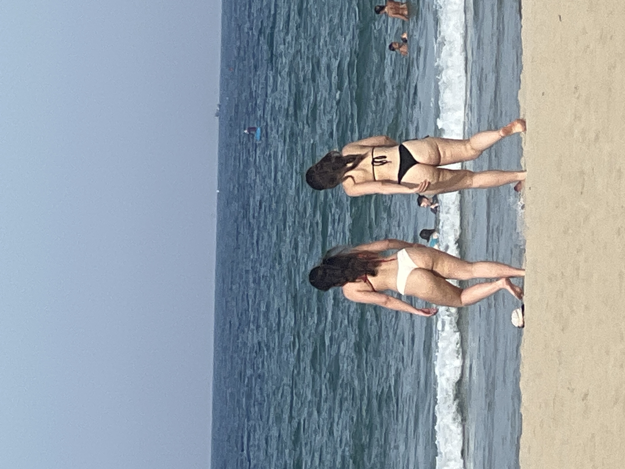 The media file shows a beach scene in Đà Nẵng, Vietnam, with the ocean and sand clearly visible. There are several people in the water and on the sand. Two women are prominently featured, both wearing bikinis. One woman in a black bikini is in the foreground, facing away from the camera with her head tilted down. Another woman in a white bikini is positioned slightly behind and to the right of the first woman, also facing away from the camera, with her legs extended towards the sand. Several other individuals are visible in the background, some wading in the water and others seemingly further out. The water is a deep blue with waves breaking near the shore, and the sand is a light beige. The sky is a clear, pale blue, suggesting it is daytime with fair weather. No text is visible in the media file. The overall impression is a casual day at the beach, with people enjoying the ocean and sunshine.