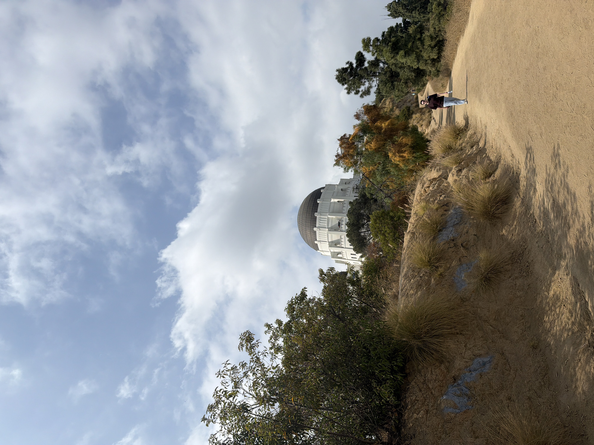 The image shows the Griffith Observatory in Los Angeles, California. The observatory is visible behind a grassy hillside with trees. A person is walking along a dirt path that runs along the top of the hill. The sky is visible in the background, with a mix of blue sky and white clouds.