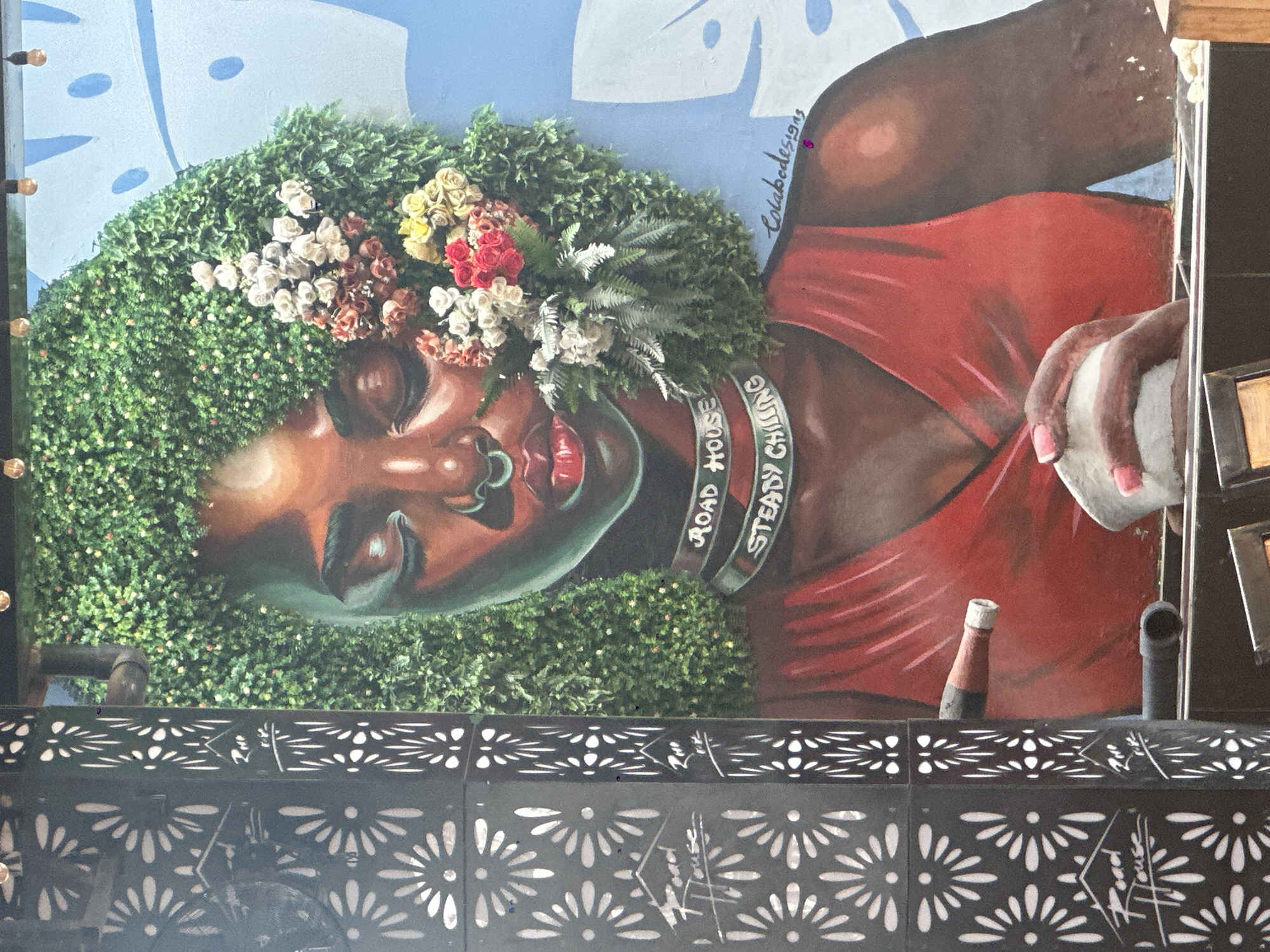 This image shows a mural of a Black woman with her eyes closed, wearing a red top and a floral-adorned green headdress. The mural is located on a wall in the city of Owerri, Nigeria. The woman's hair is made of artificial greenery, and she has a nose ring. She is wearing a choker-style necklace that reads "ROAD HOUSE STEADY CHILLING". Her hands are holding a white object with pink-tipped fingernails. A bottle is shown in the foreground, pointing towards the woman. To the left of the mural is a black metal screen with a repeating flower pattern. The background of the mural is a light blue with abstract white shapes. A signature that reads "Colabodesigns" is visible in the lower right corner of the mural.
