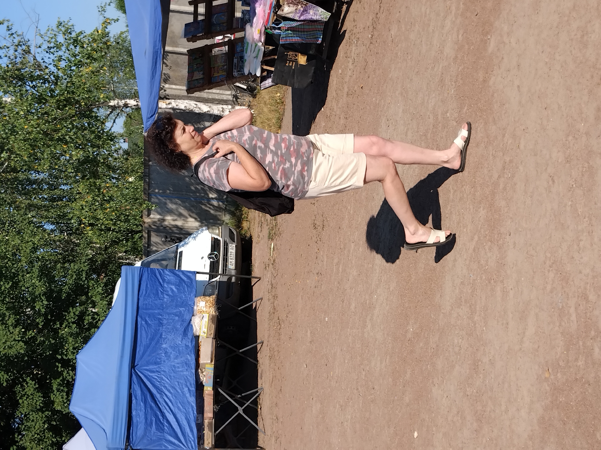 A woman with curly brown hair, wearing a camouflage t-shirt and cream-colored shorts, walks rightward across a dirt lot. She is carrying a black backpack and her right hand is near her face. She is wearing white slide sandals. A shadow of her is cast on the ground to her left. In the background, a blue tent shelters a table laden with boxed goods. To the right of the tent, a white van is parked. Further to the right, a fence separates the lot from trees and buildings. Another stall with shelves displays books and other items. The scene appears to be a market in Irshansk, Ukraine.